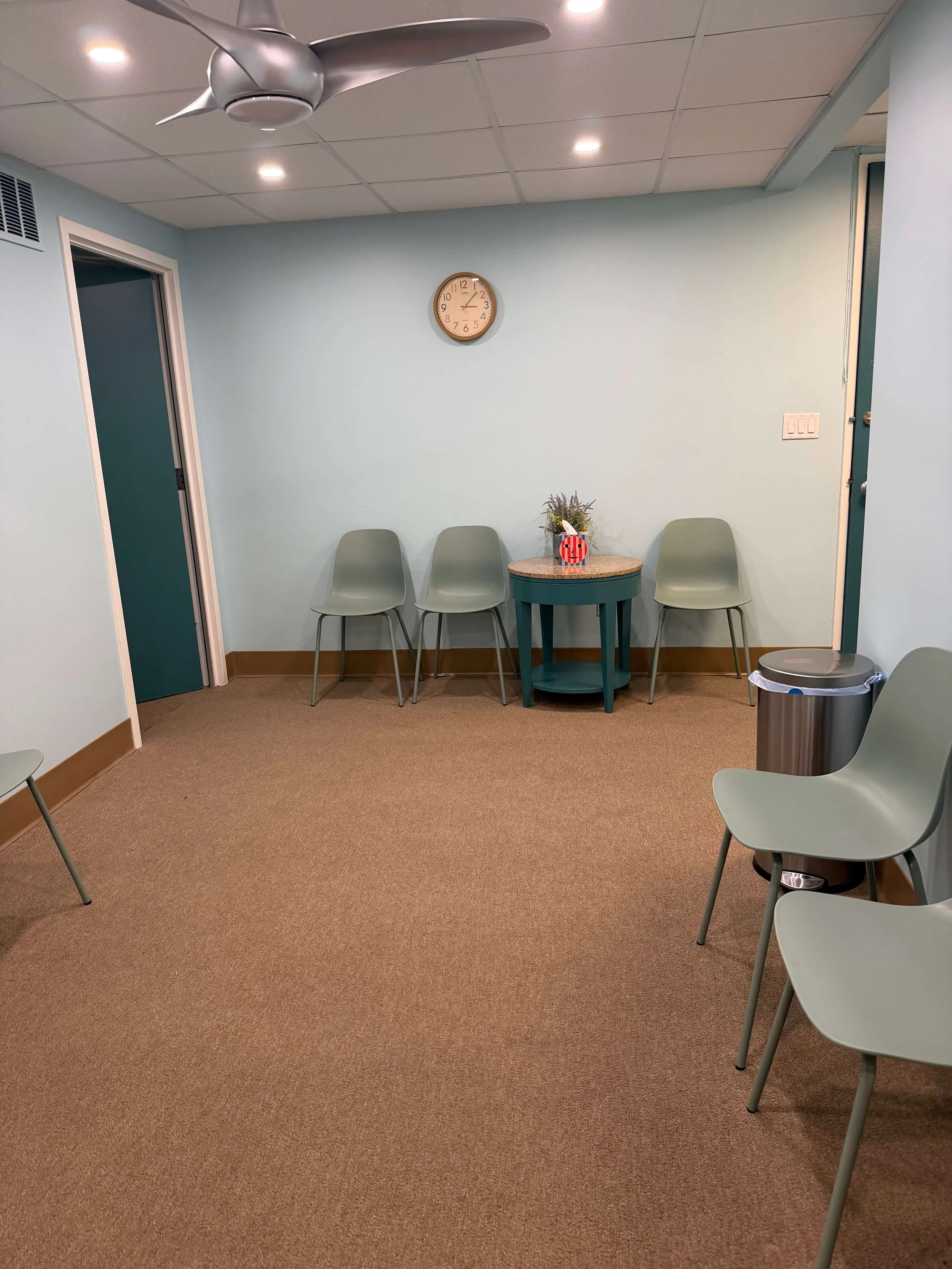 A waiting area with four light green chairs, a small round table with a potted plant and gift bag, a trash can, a wall clock showing 1:10, light blue walls, an open door, and a ceiling fan with lighting.