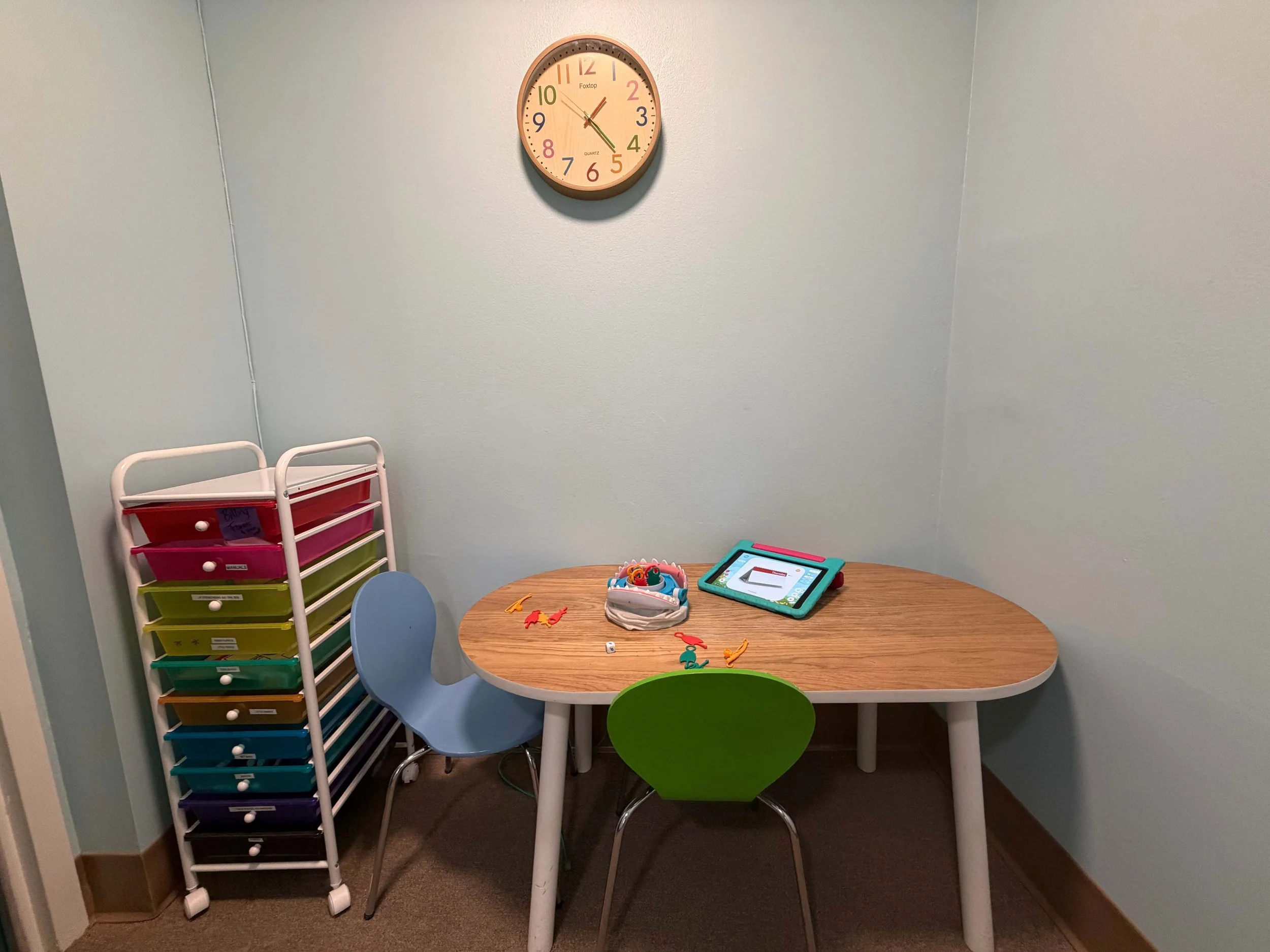 A small playroom corner with a round table, two colored chairs, a plastic storage unit with drawers, and children's toys on the table. There is a clock on the wall showing approximately 12:10.