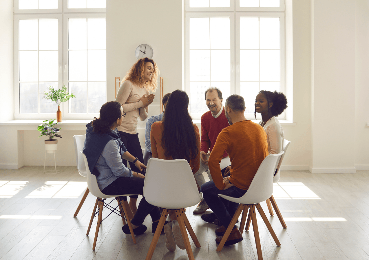 A diverse group of people sitting in a circle and engaging in a lively discussion or meeting in a bright, well-lit room with large windows and minimal decor.