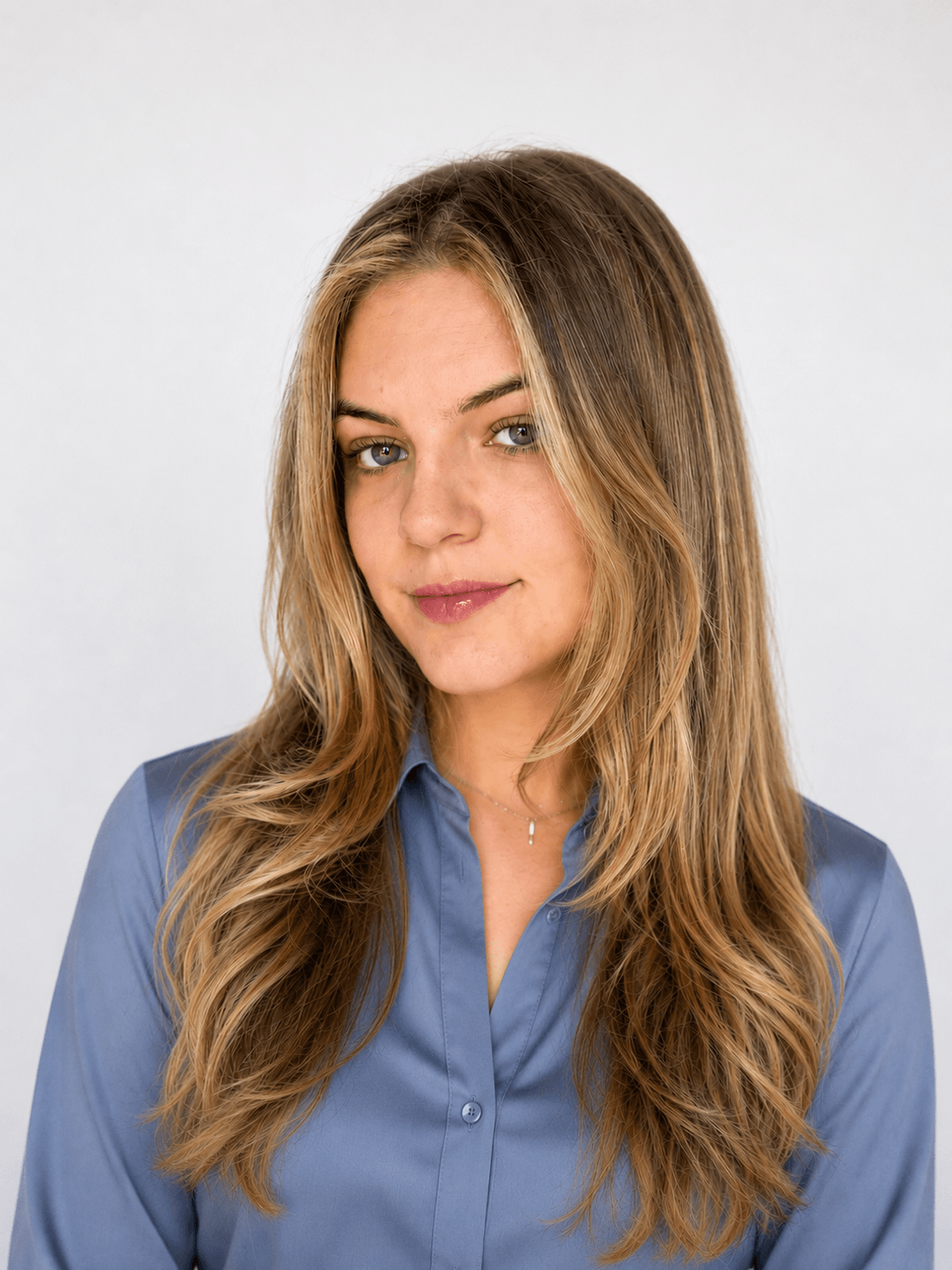 A young woman with long, wavy blonde hair wearing a blue button-up shirt, looking at the camera with a slight smile, against a plain white background.