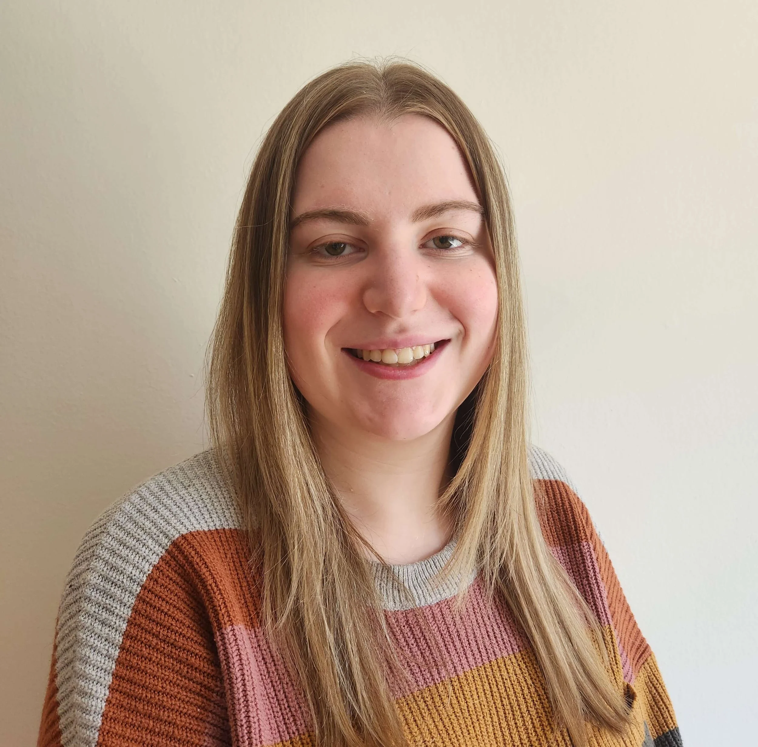 Close-up of a young woman with long blonde hair, smiling and wearing a multicolored striped sweater, standing against a plain light-colored wall.