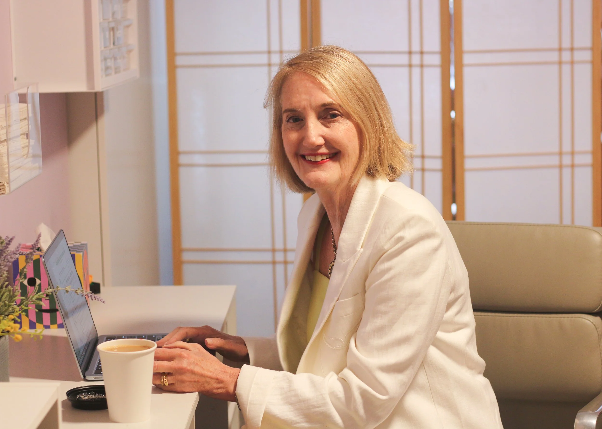 A woman with blonde hair and a light-colored blazer working on a laptop at a desk, with a cup of coffee in front of her, in an office or workspace setting.