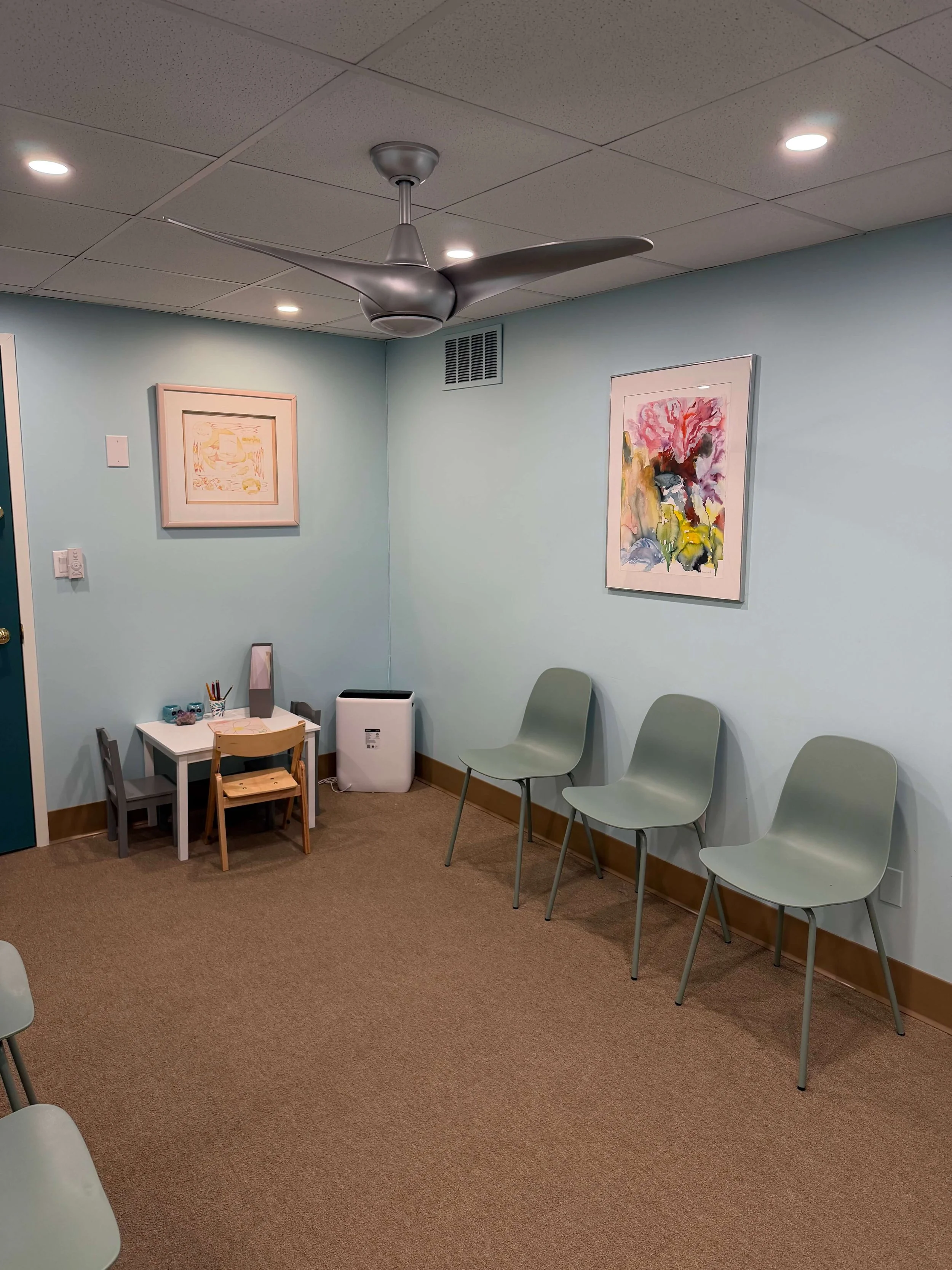 A waiting area with four green chairs in a row against a light blue wall, two watercolor paintings on the wall, a small table holding art supplies, an air purifier, and a ceiling fan.