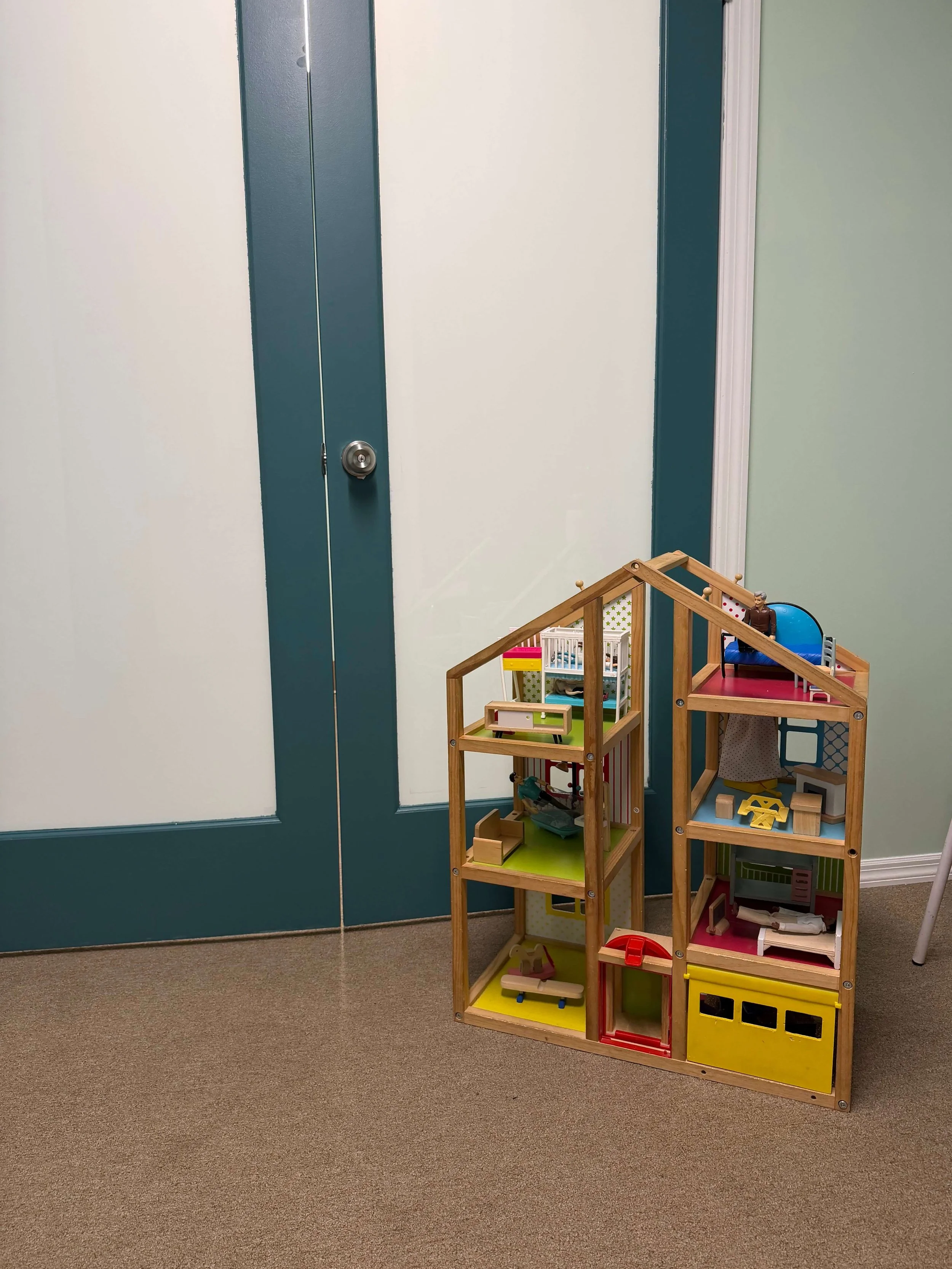 A wooden dollhouse with multiple rooms and furniture, placed against a teal door and light green wall in a room with beige carpet.