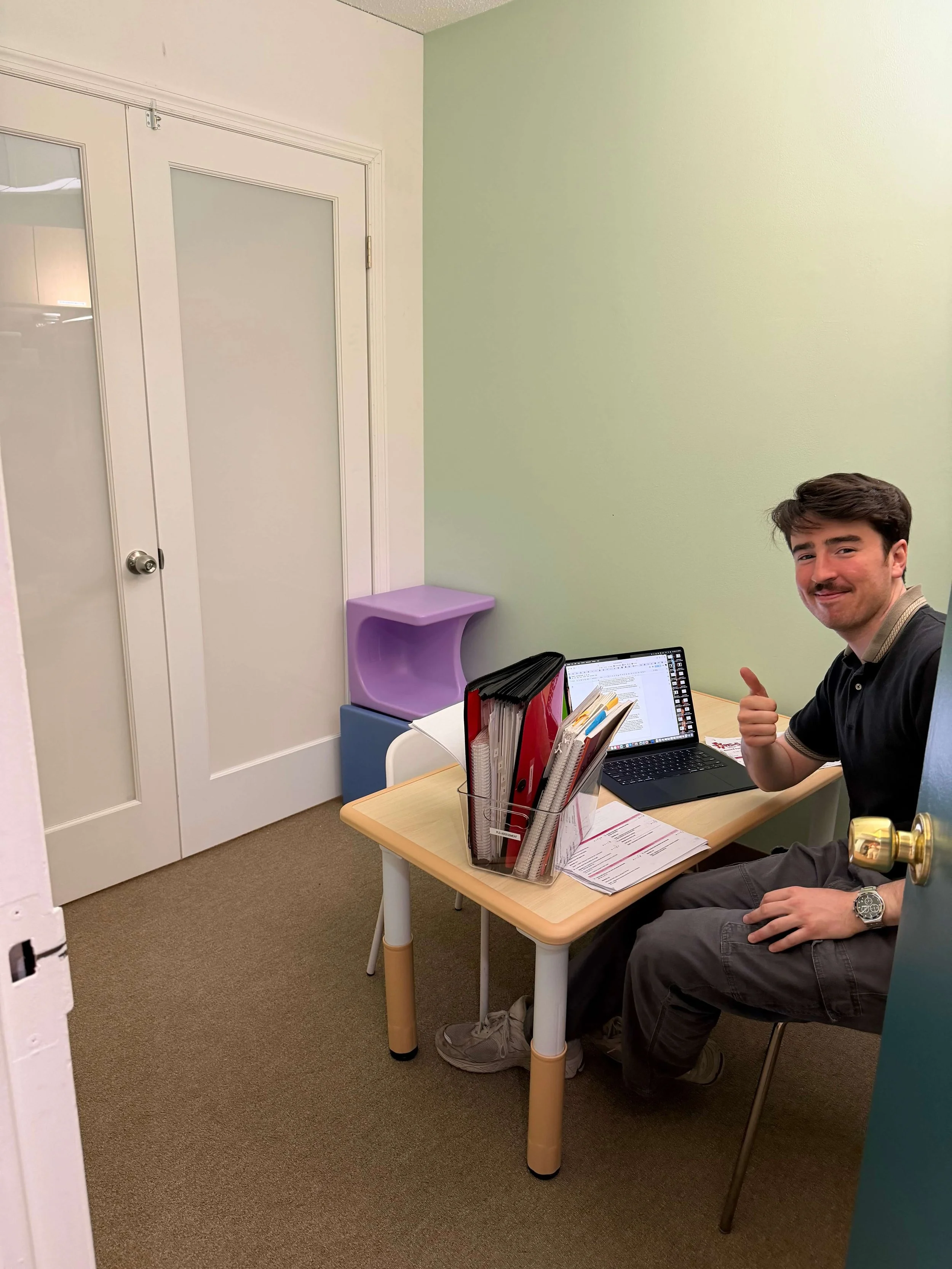 A man sitting at a small table with a laptop and organized folders, giving a thumbs-up gesture in a small, plain room.