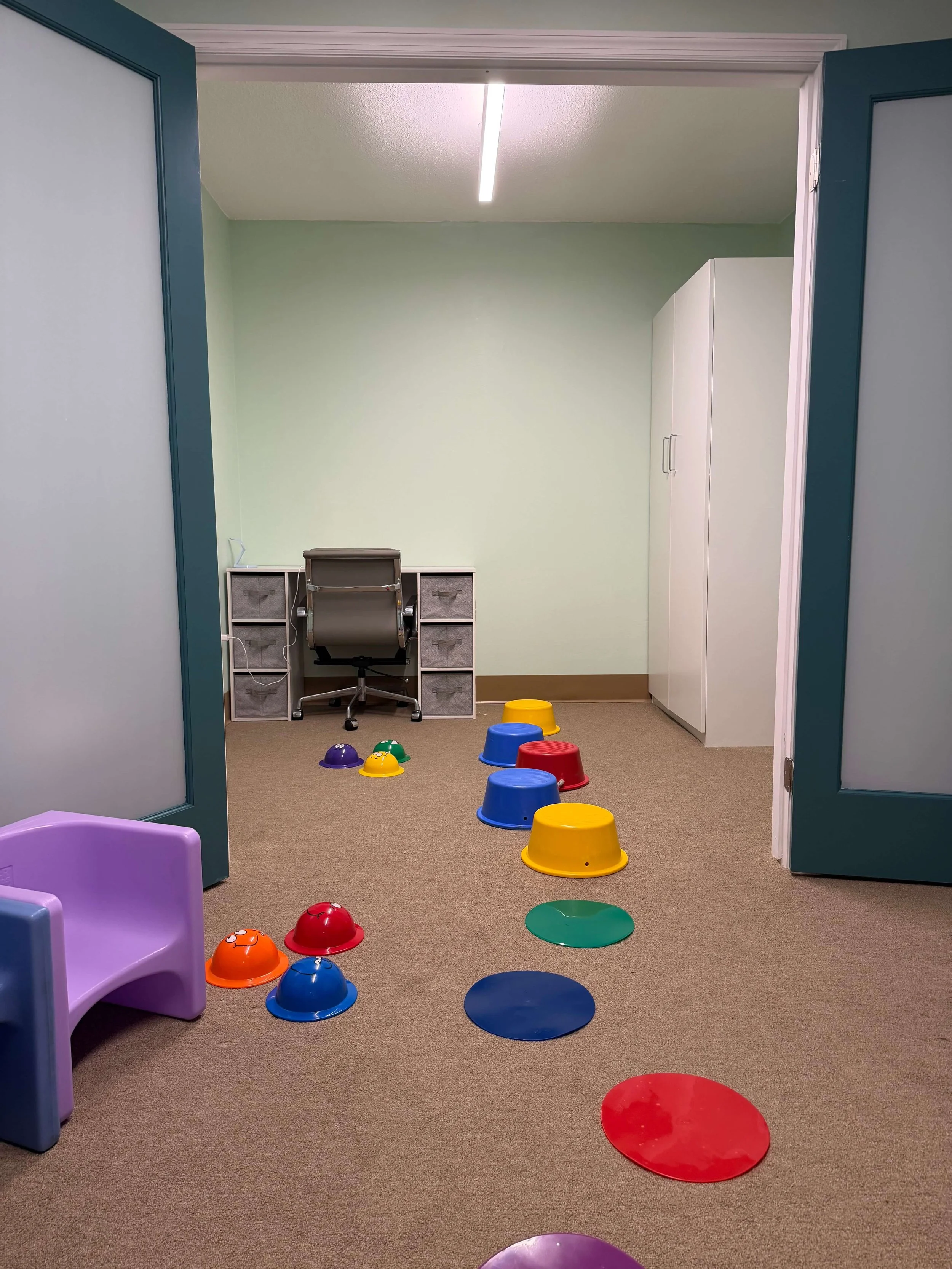 A playroom with small colorful plastic chairs and bowls arranged on a beige carpeted floor, a desk with a chair and storage cubes, a white cabinet, and a light green wall with a ceiling fluorescent light.