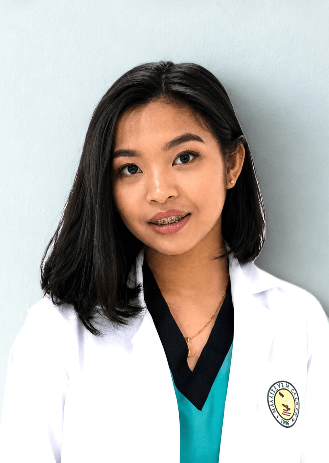 A young woman with shoulder-length black hair, wearing a white medical coat with a patch, a black and teal scrubs, a gold necklace, and braces, smiling and standing against a light-colored wall.