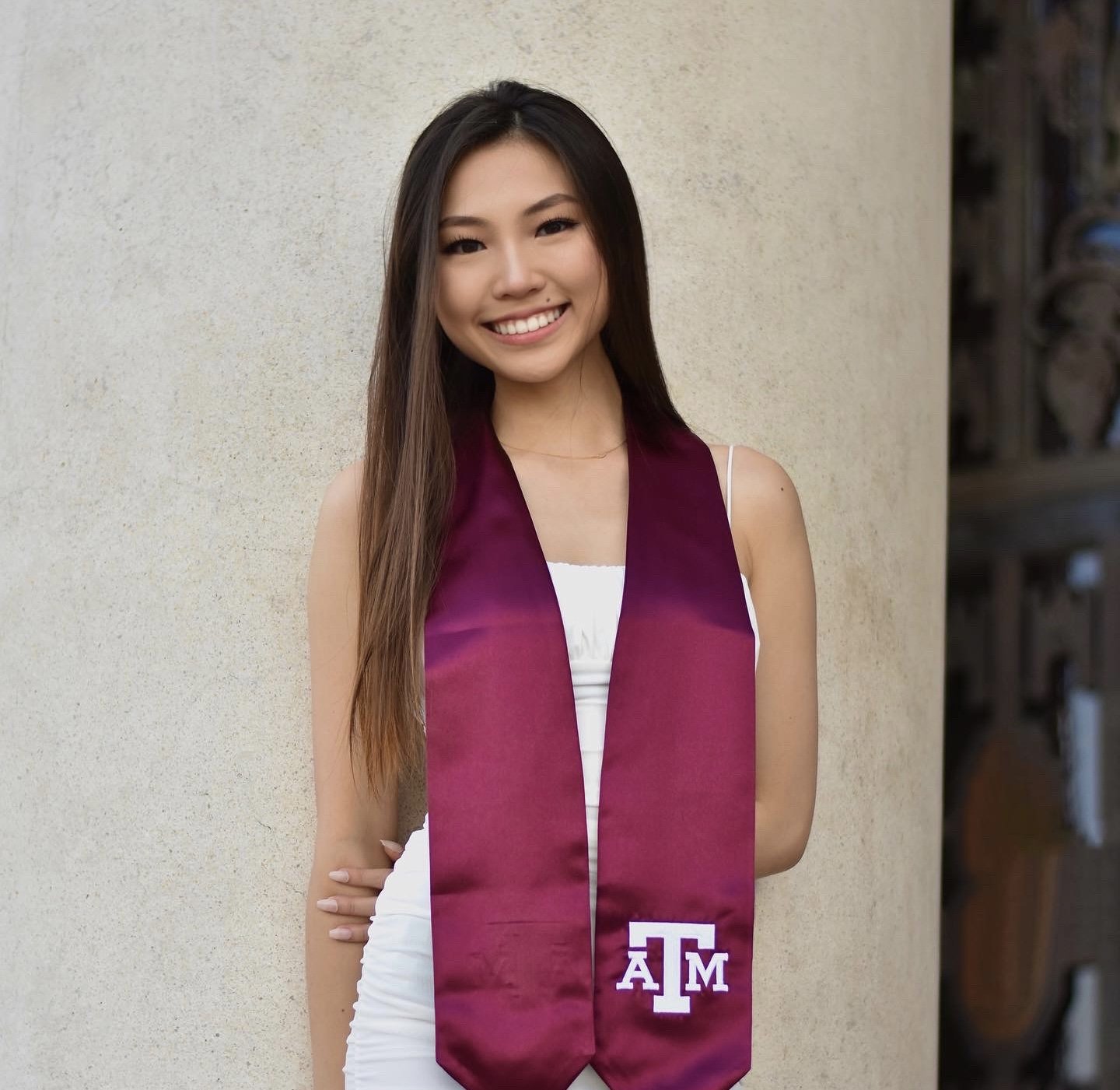 Smiling young woman in white dress wearing a maroon graduation stole with Texas A&M University logo