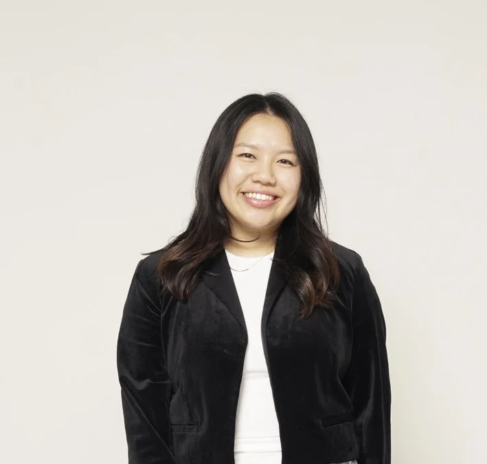 A smiling woman with long dark hair wearing a black blazer and white top standing against a plain light-colored background.