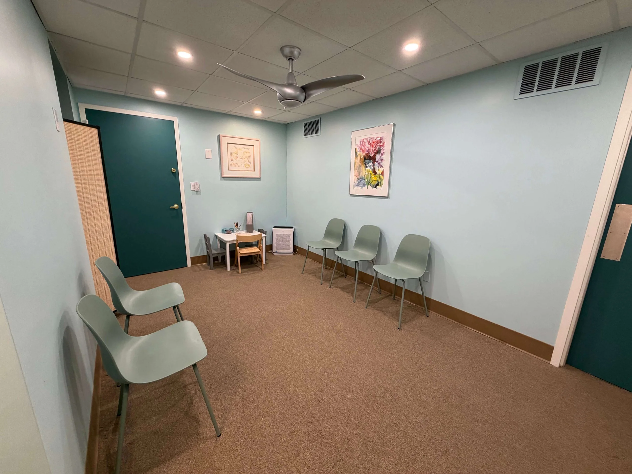 An empty waiting room with light green chairs arranged along the walls, framed artwork on the walls, a small table with chairs, a small heater, and ceiling vents.