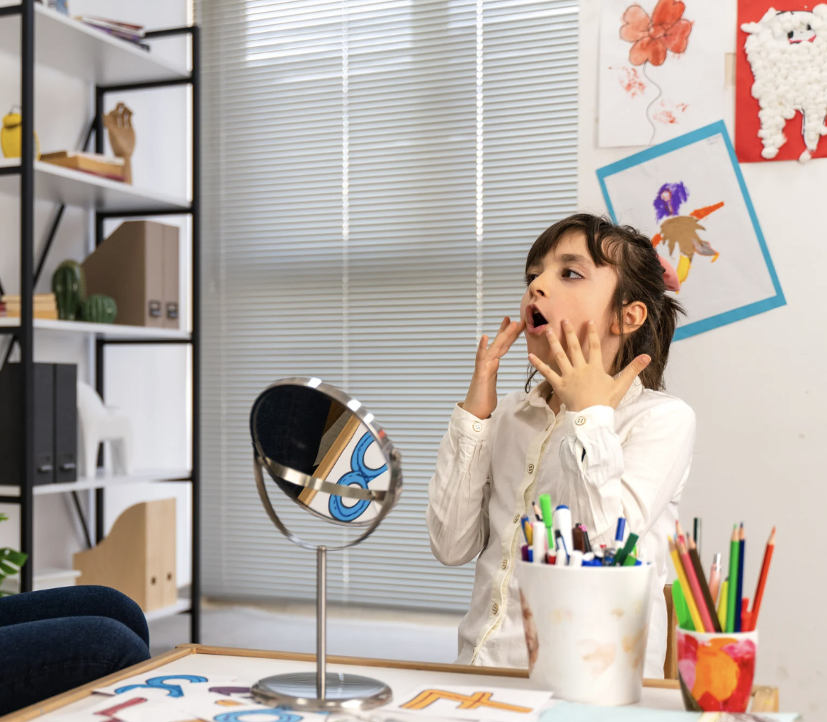 A young girl with dark hair, wearing a white shirt, stands in front of a mirror, making a surprised face with her hands on her cheeks in a room decorated with colorful children's artwork and art supplies.