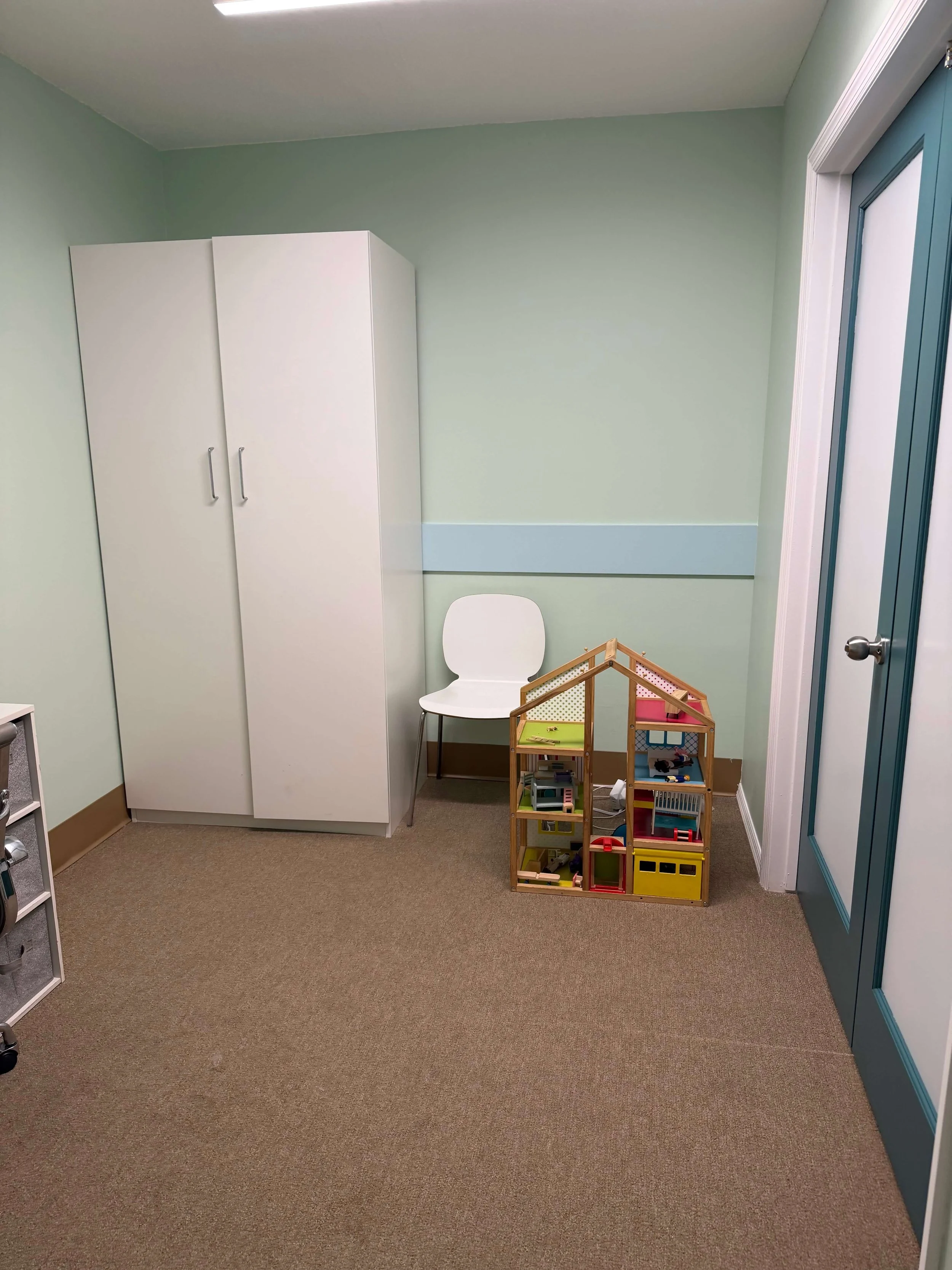 A small room with green walls, a white cabinet, a white chair, a wooden dollhouse, and a blue door.
