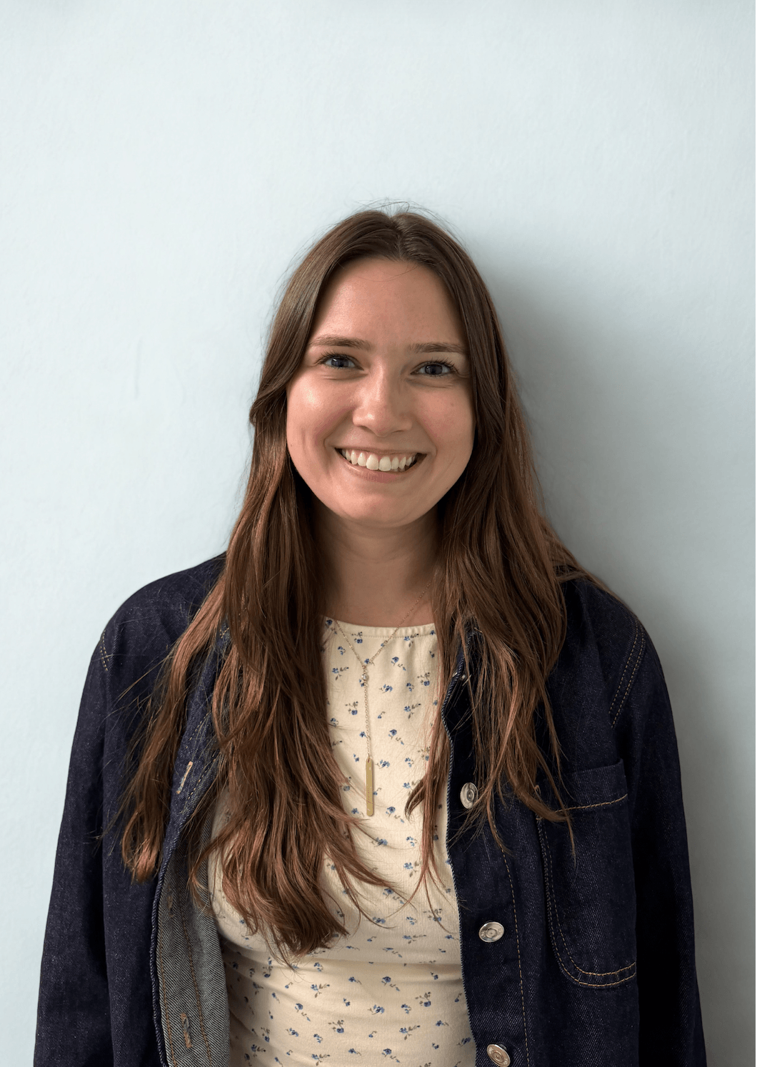 Young woman with long wavy brown hair smiling, wearing a dark denim jacket over a light-colored patterned shirt, standing against a plain light-colored wall.