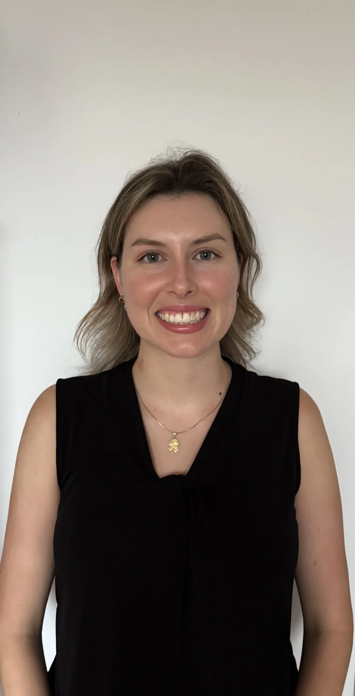 A woman with shoulder-length wavy blonde hair, wearing a black sleeveless top, a gold necklace with a pendant, and small earrings, smiling in front of a plain white wall.