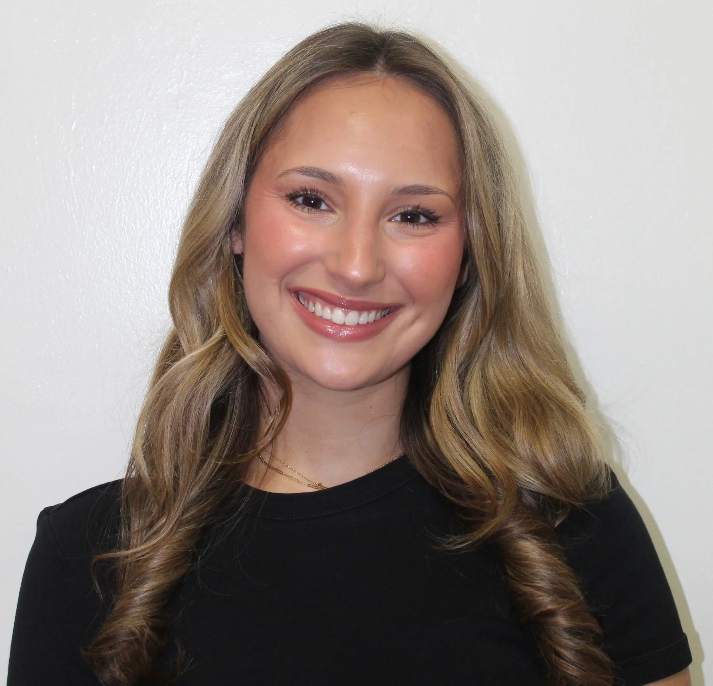 Close-up photo of a woman smiling, with long wavy light brown hair, wearing a black top, standing against a white wall.