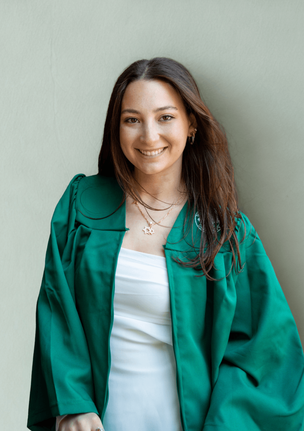A young woman with long brown hair smiling, wearing a green jacket over a white top and layered necklaces, standing against a light green wall.