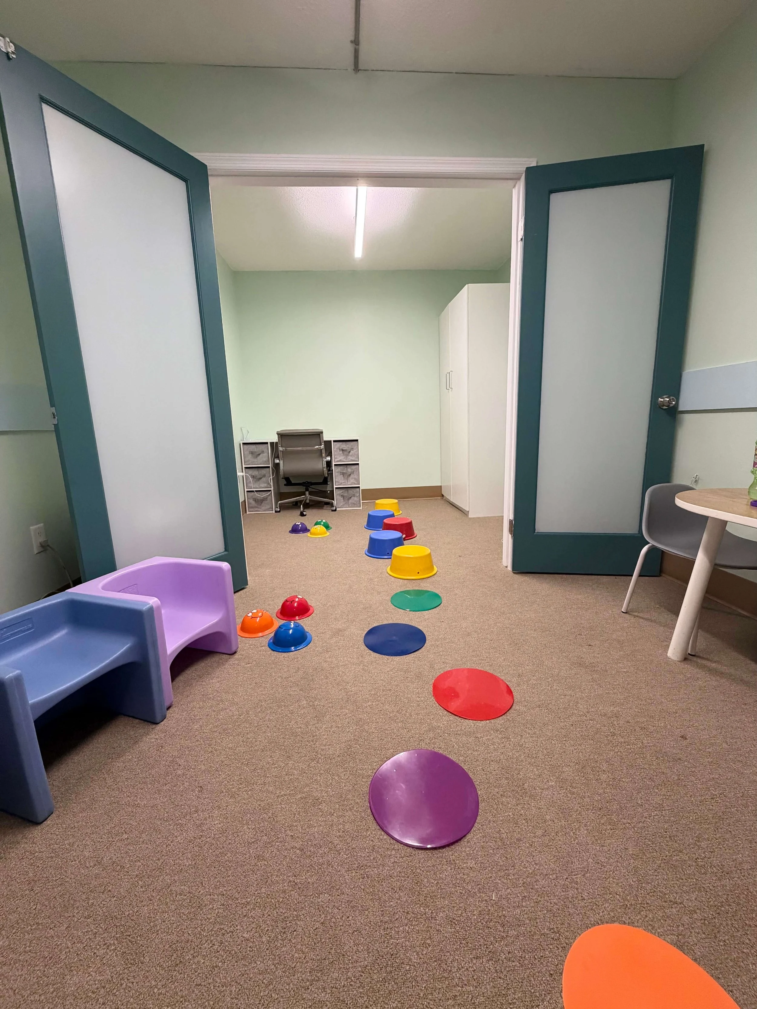 Colorful stepping stones arranged in a path on a beige carpet in a children’s playroom. The room has green walls, a white cabinet, a desk with a chair, and some small furniture pieces.