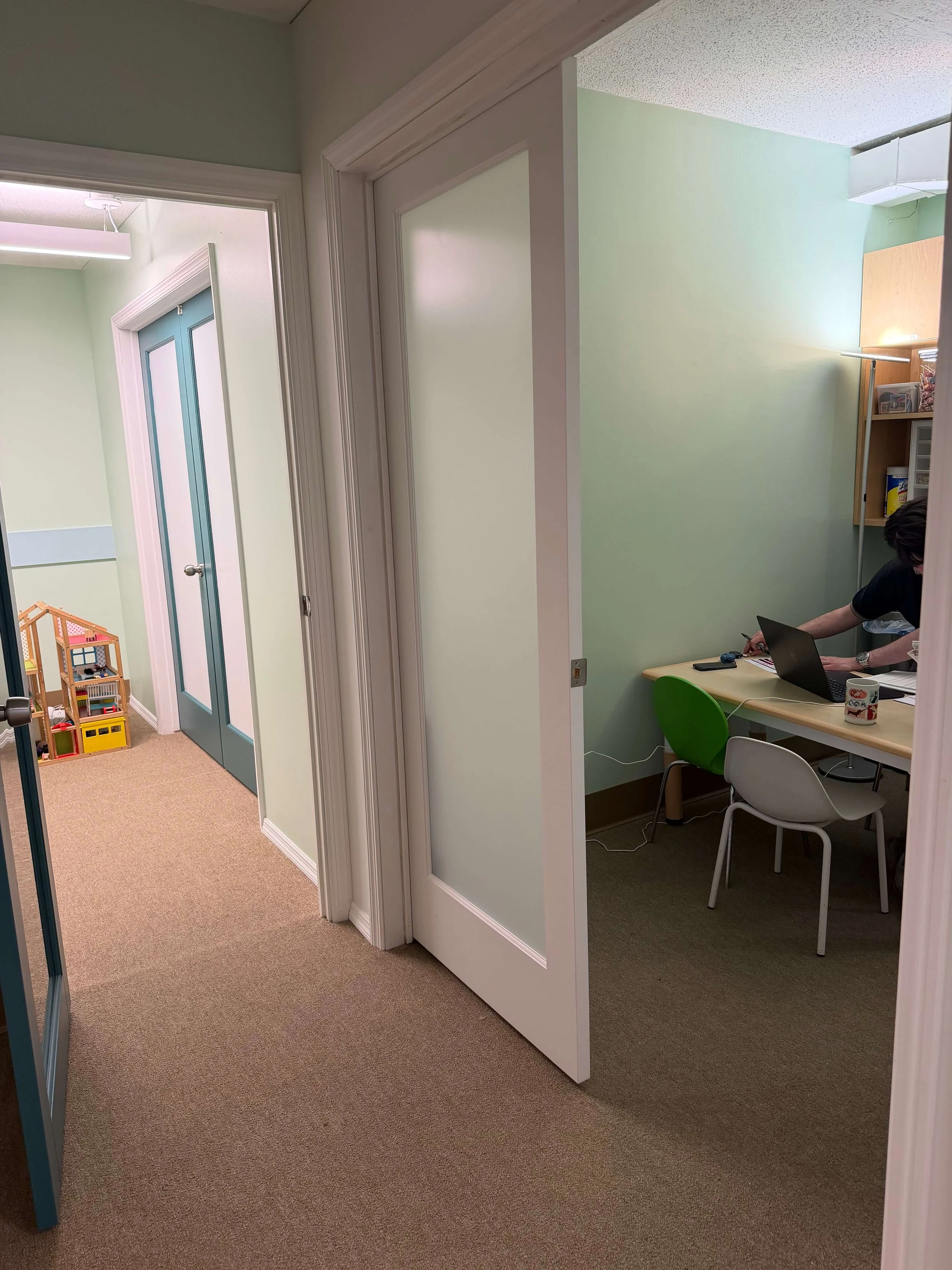 Office hallway with beige carpet, a green wall, and a small meeting room with a person sitting at a desk using a laptop.