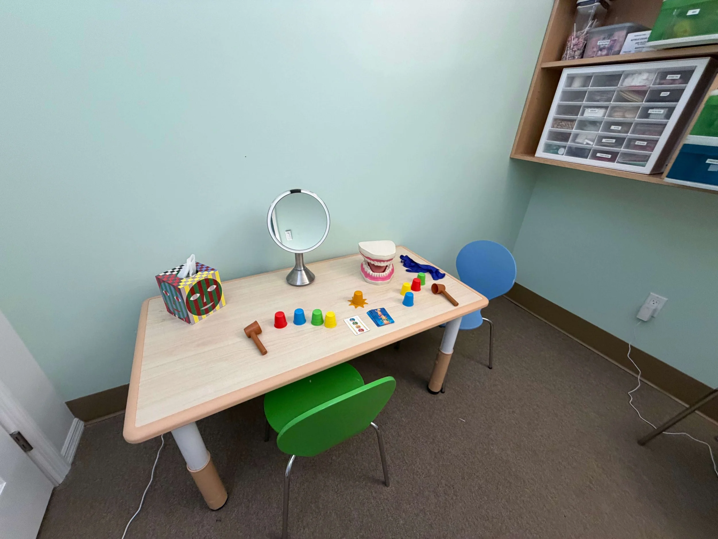 A small wooden table with educational toys and a giant plastic mouth model. The table has colorful cups, a yellow toy, and a deck of cards. Behind the table is a blue chair, and the room has a mint green wall with a white electrical outlet and a white ring mirror hanging from a stand on the table. There is a wooden shelf against the wall holding a box of small drawers and other storage containers.