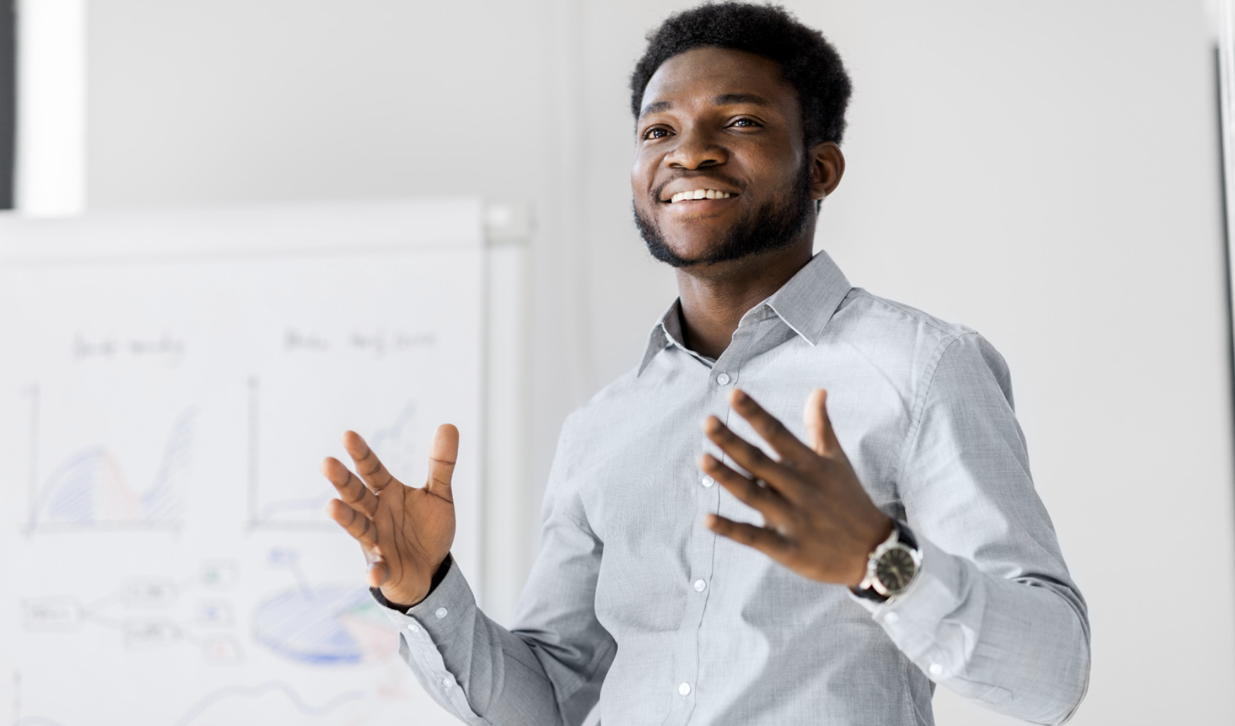 A young man in a light gray dress shirt giving a presentation in front of a whiteboard with charts and graphs, smiling and gesturing with his hands.