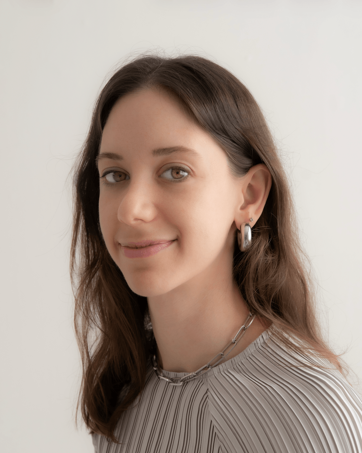 A young woman with brown hair, hoop earrings, and a silver chain necklace, smiling slightly, wearing a striped top, standing against a plain light background.