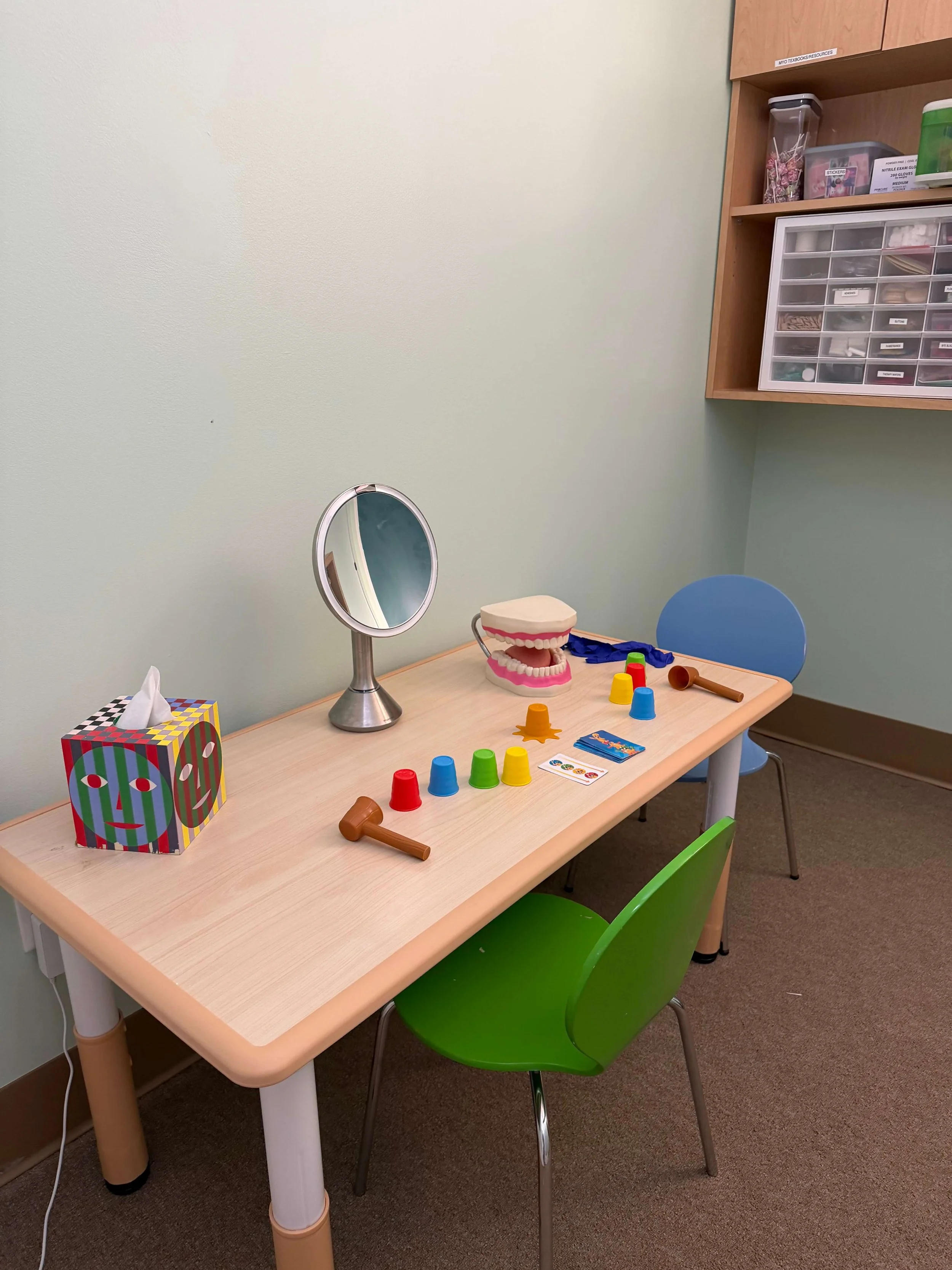 Dental examination room with a table holding a dental model, colorful cups, a mirror, and dental tools. There are green and blue chairs around the table. A wall-mounted cabinet in the background contains containers with dental supplies.