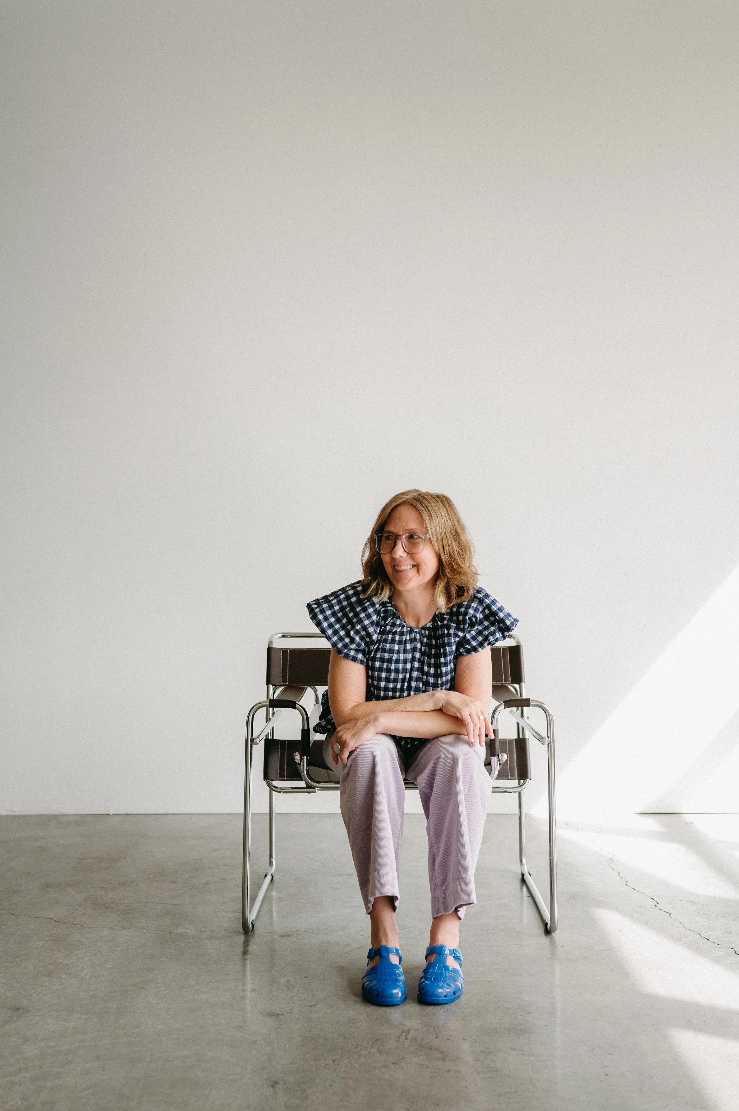 female coach sits in chair looking off to her right, wearing a blue checked shirt and lilac corduroy pants
