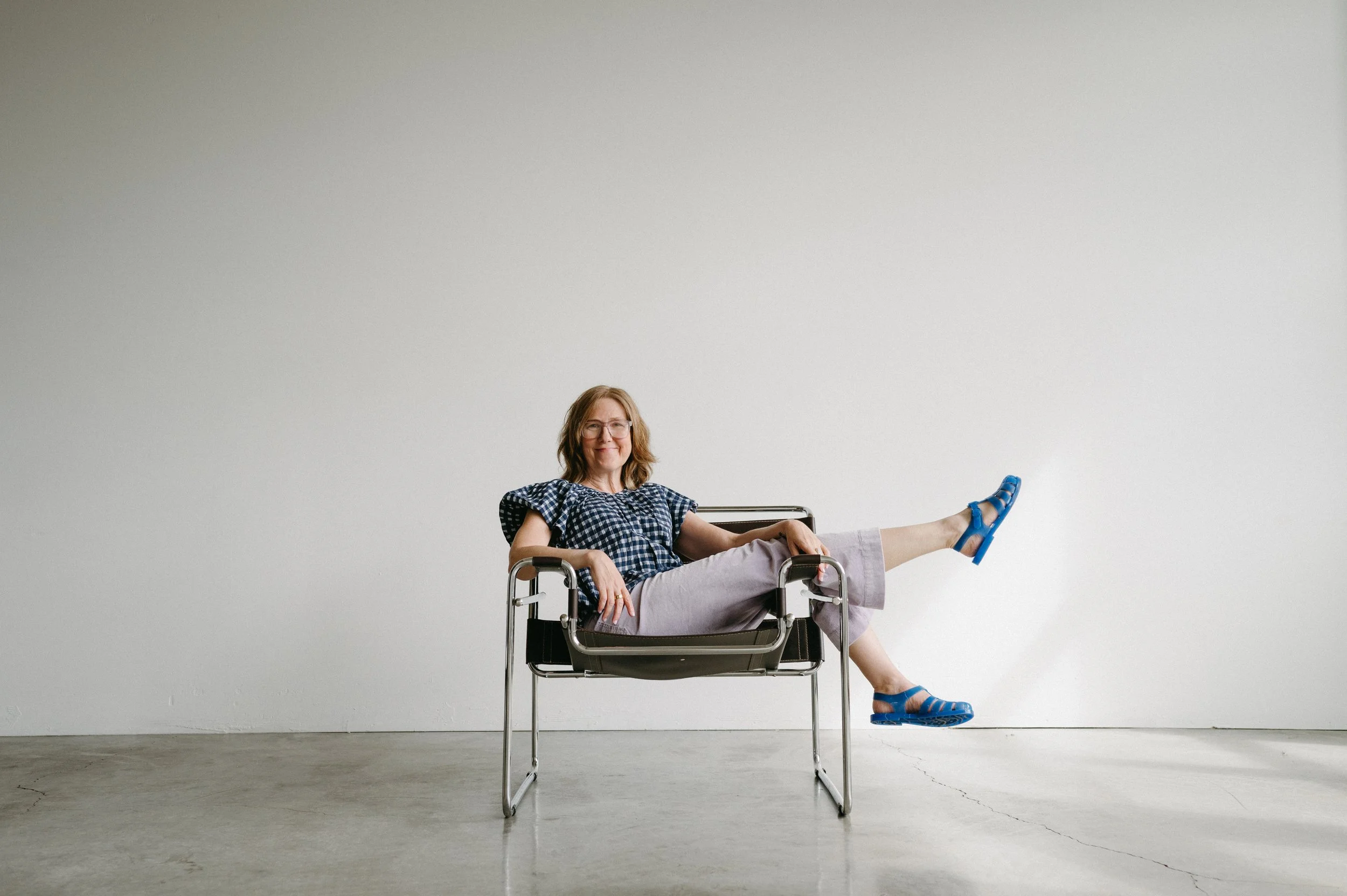 female coach sits sideways in her chair with her legs over the arm rest, one of them kicks playfully out. she is wearing glasses, a blue gingham checked shirt and lilac pants