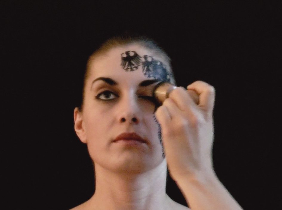 Selma Alacam Different Conditioning - Videostill: Die Künstlerin drückt sich mit einem Stempel einen schwarzen Bundesadler ins Gesicht