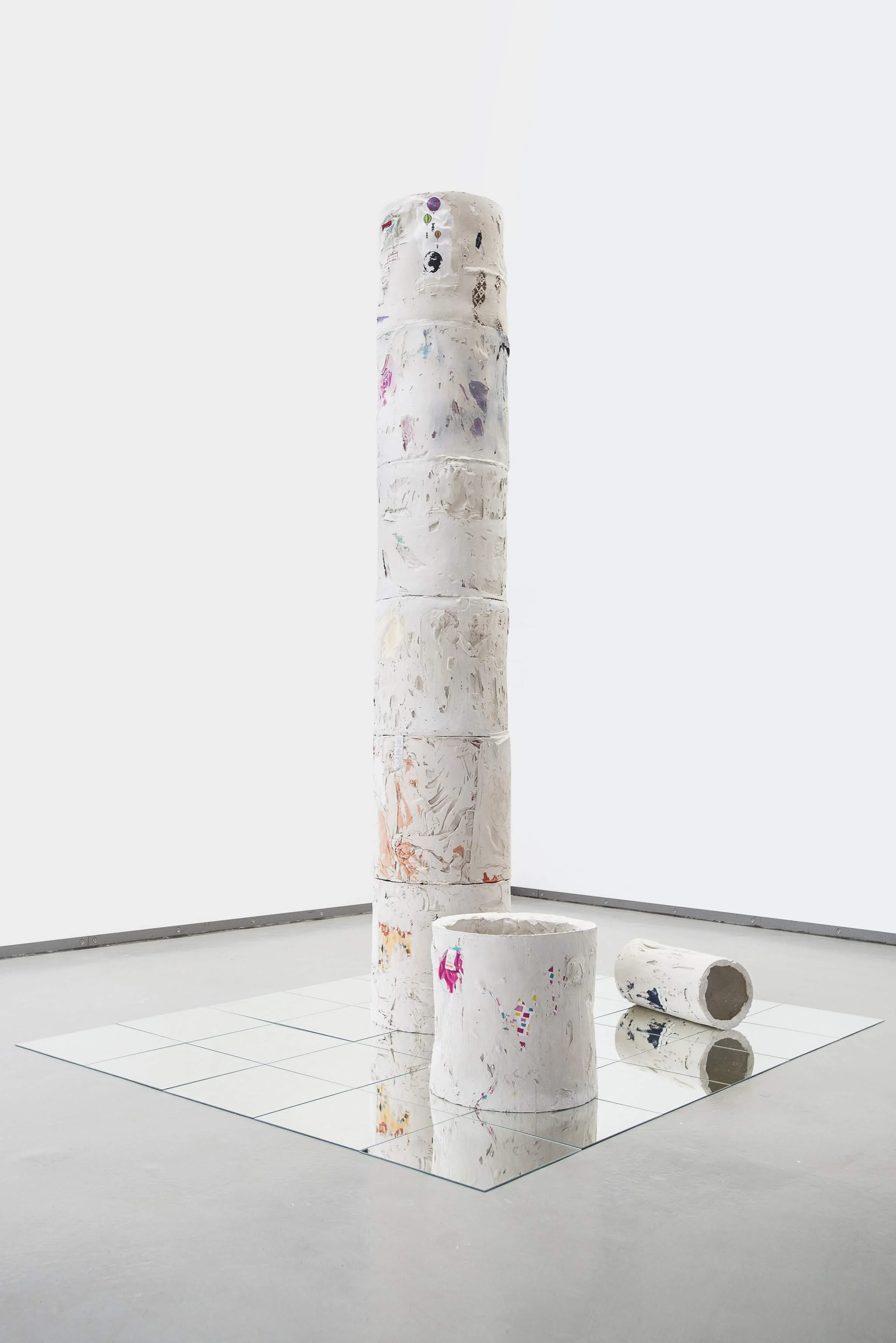 1 hohe Säule aus 6 Elementen und ein einzelnes Säulenelement stehen auf einer Spiegelfläche, an der Seite der Fläche liegt ein schmaleres Säulenelement. Aus der weißen Porzellanmasse der Hohlkörper ragen bunte Flecken der Stoffelemente heraus
