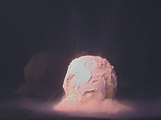 Sylvia Henze, ZEIT|EN, 2001 Videostill - eine sich in Wasser auflösende Porzellankugel vor dunkle. Hintergrund. Weiße Schlieren und Porzellanstaub, der sich am Boden um die Kugel ablagert. 