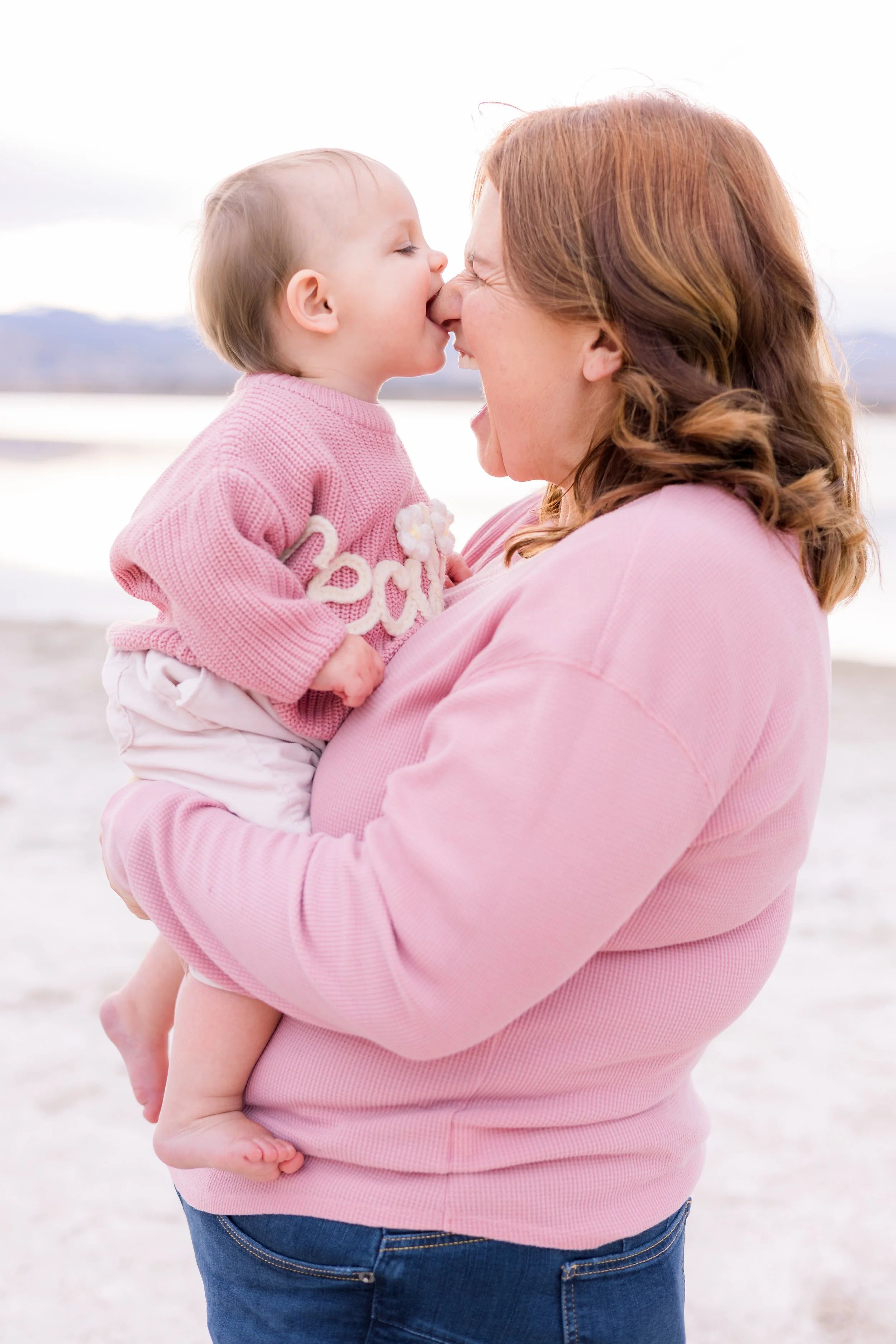 A woman holding a young girl outdoors near water, both wearing pink clothing. The girl is kissing the woman on the nose.