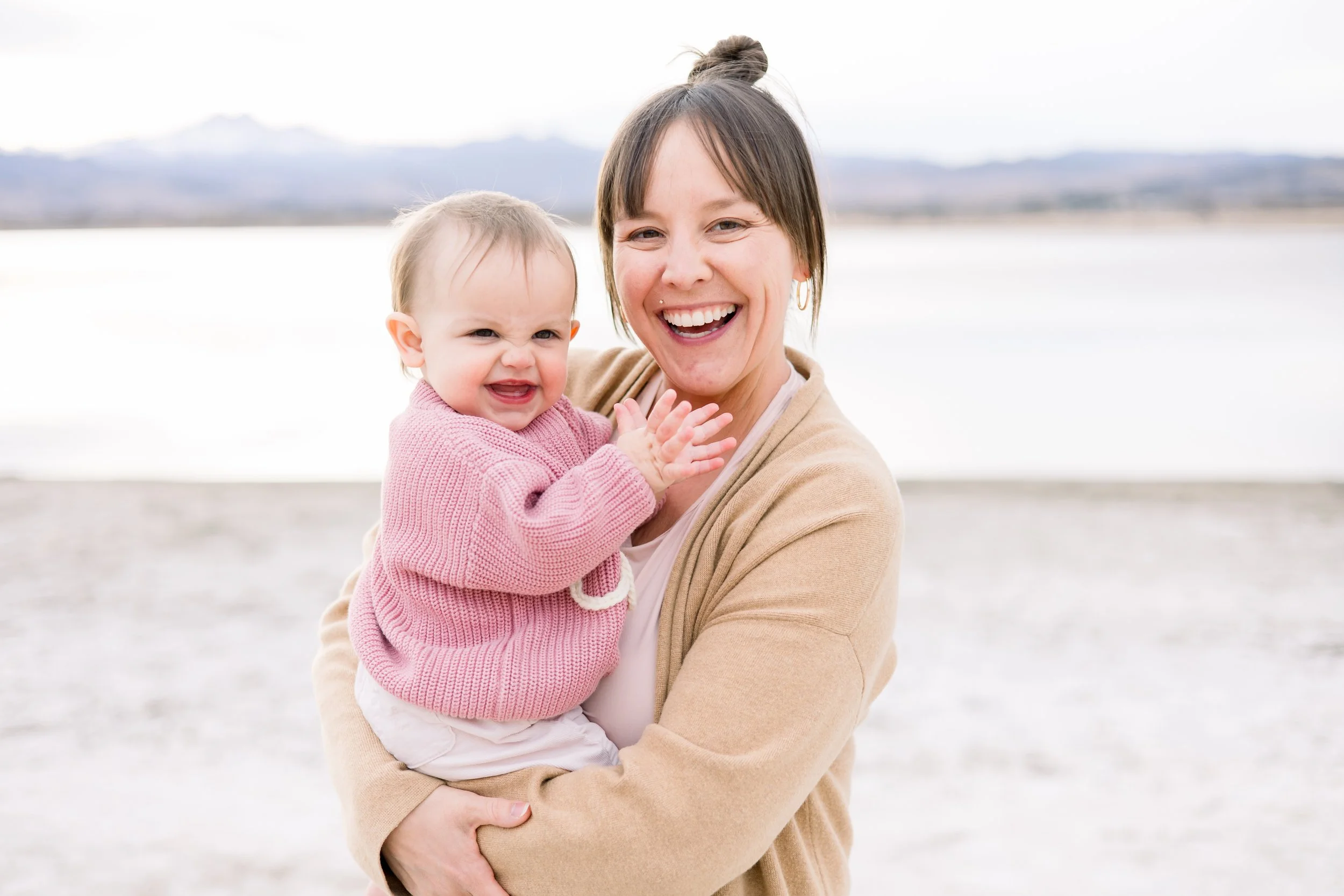 A woman holding a happy baby girl outdoors near a body of water, with mountains in the background, both smiling.