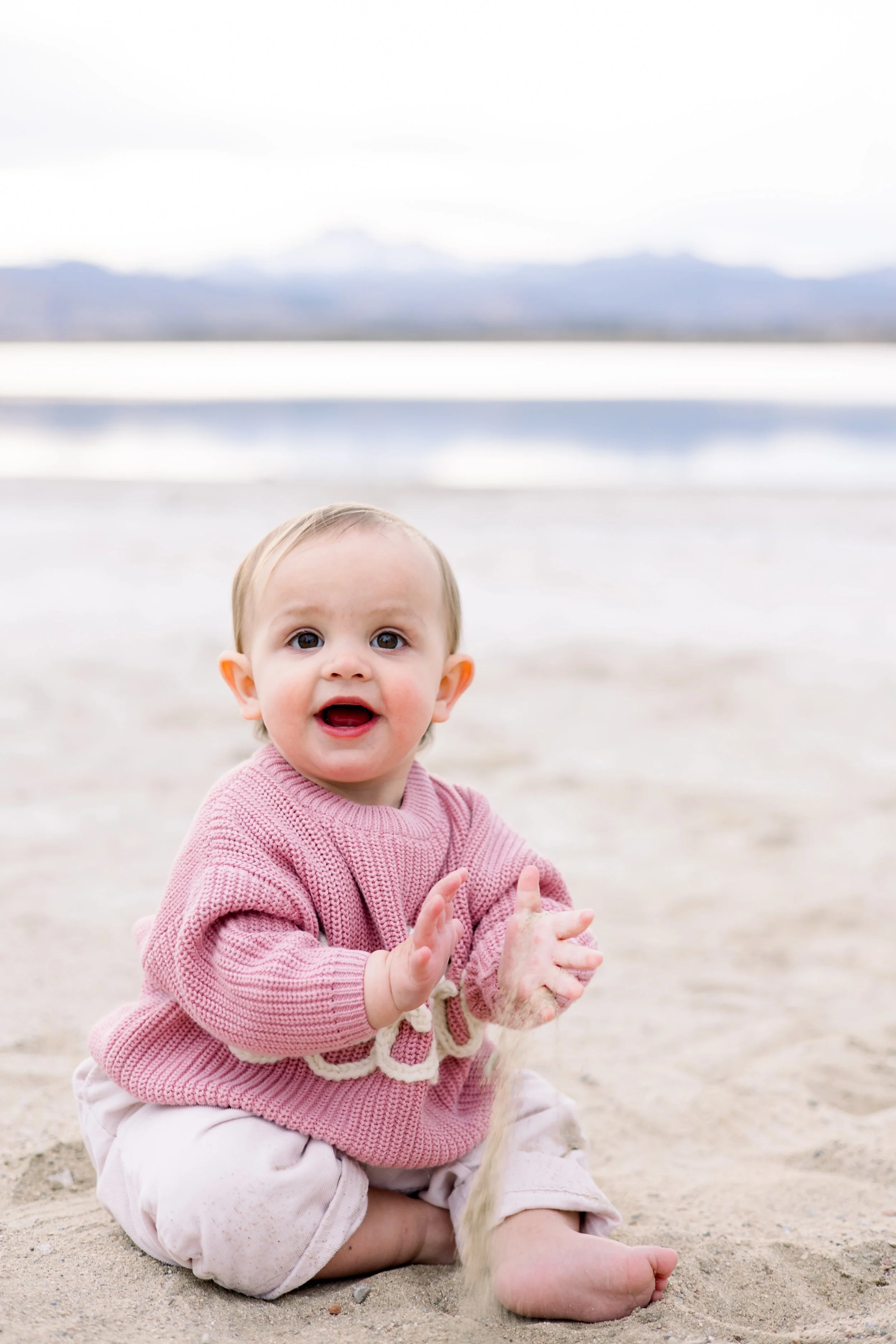 A smiling toddler girl sitting on sandy beach with water and mountains in background, wearing a pink sweater and beige pants.