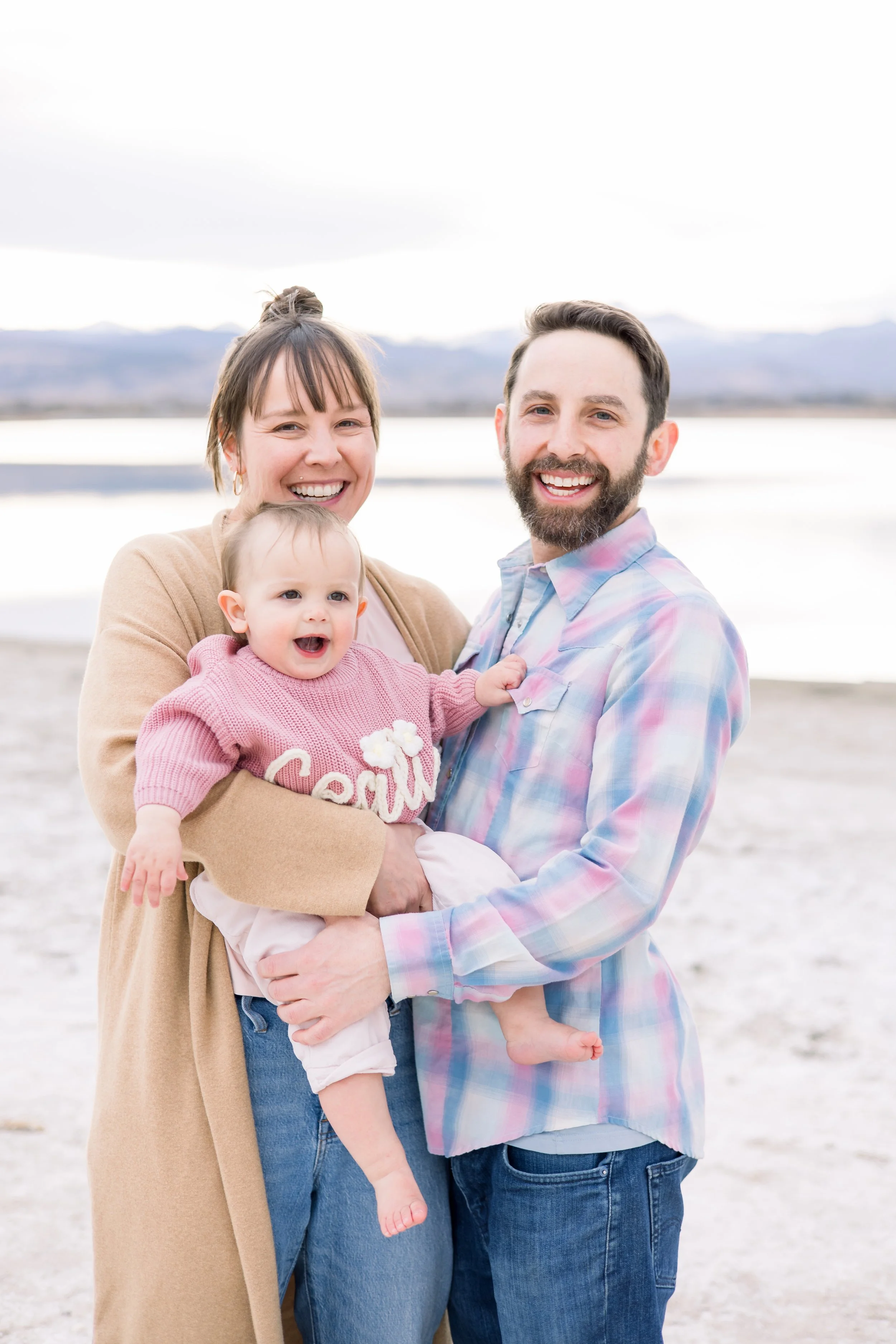 A happy family with a mother, father, and baby girl smiling at the camera on a beach with mountains and water in the background.