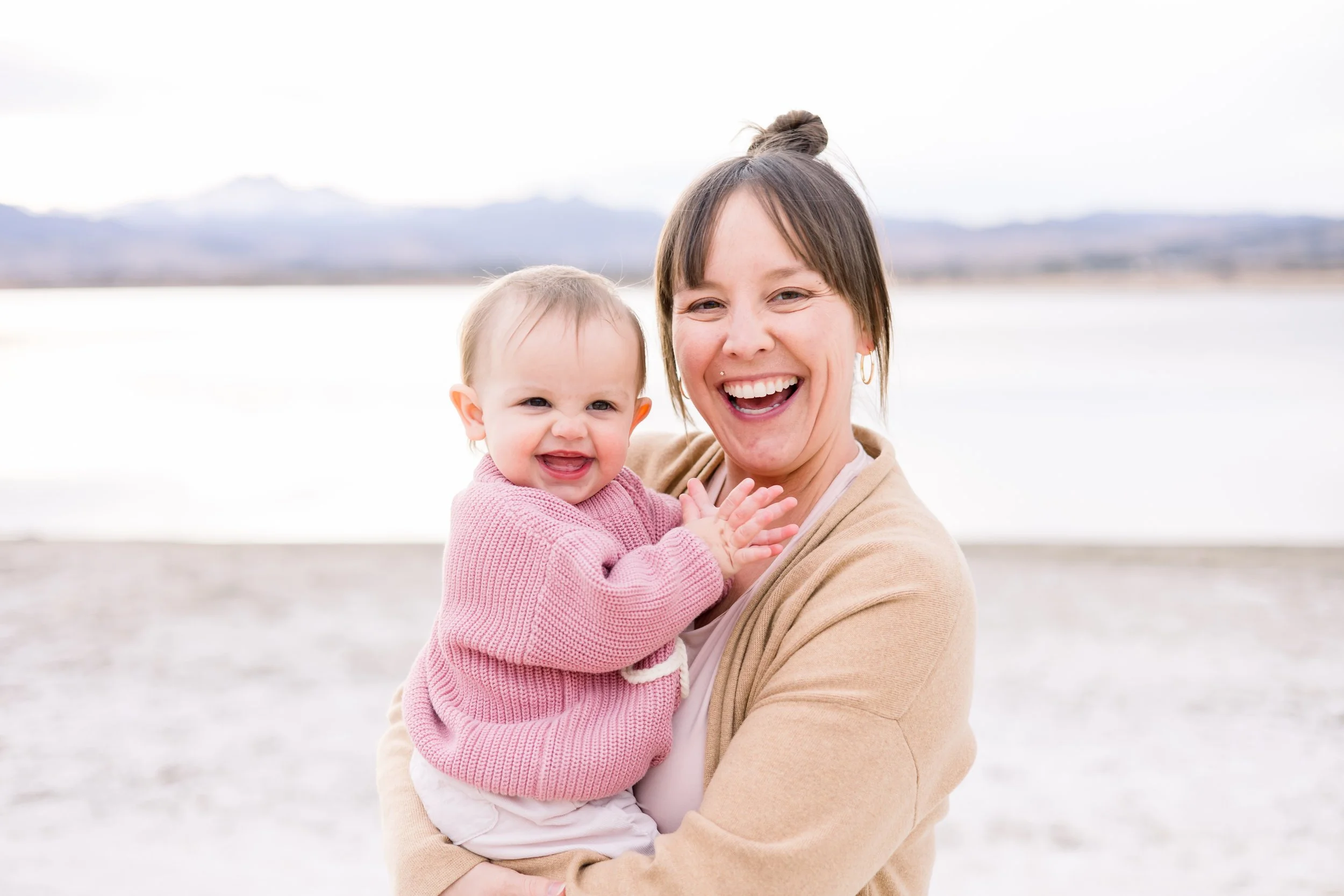 Smile of a woman holding a happy baby girl near a body of water with mountains in the background.