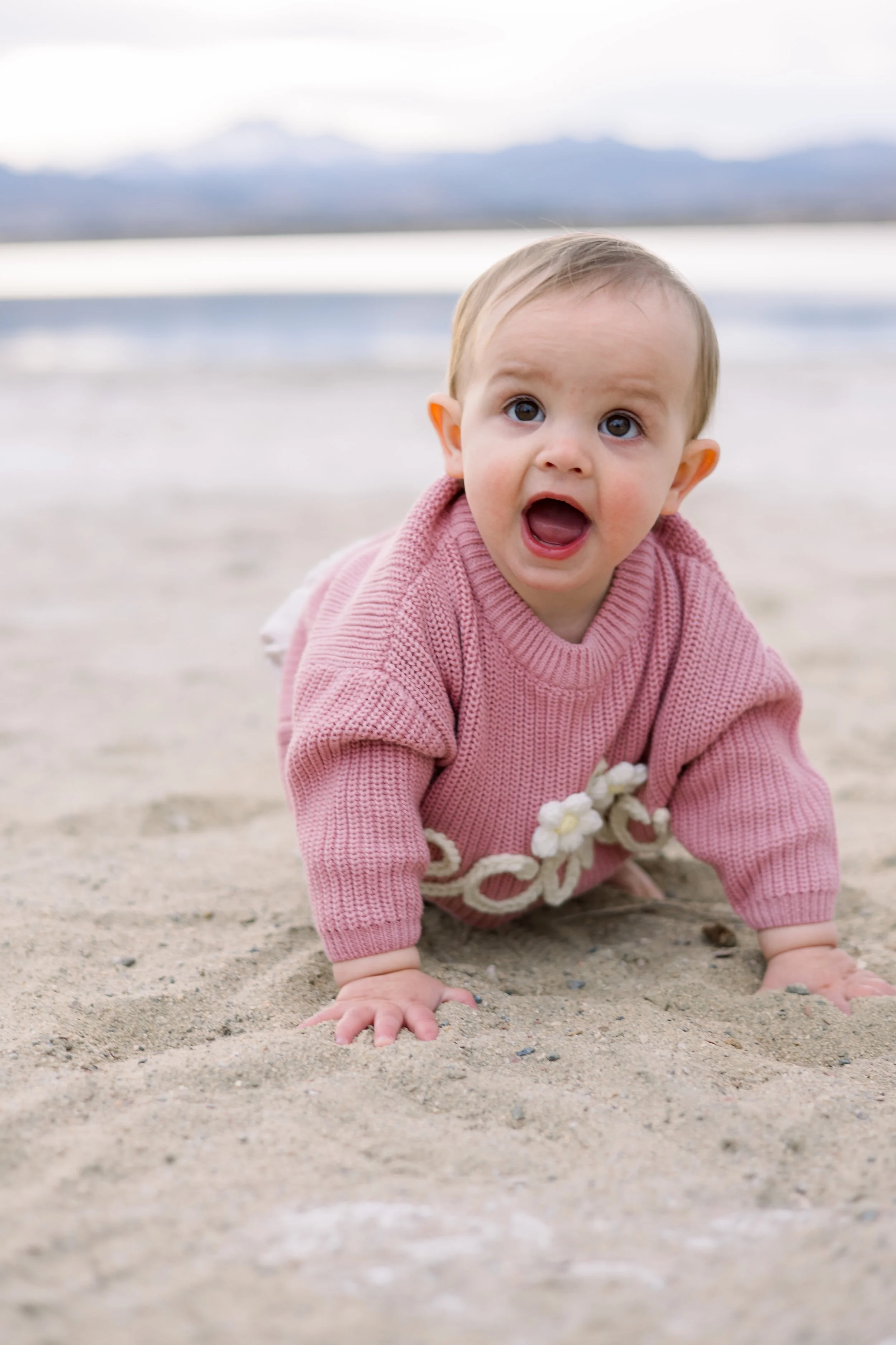 A baby crawling on the sandy beach with a surprised expression, wearing a pink sweater with white decorative elements.