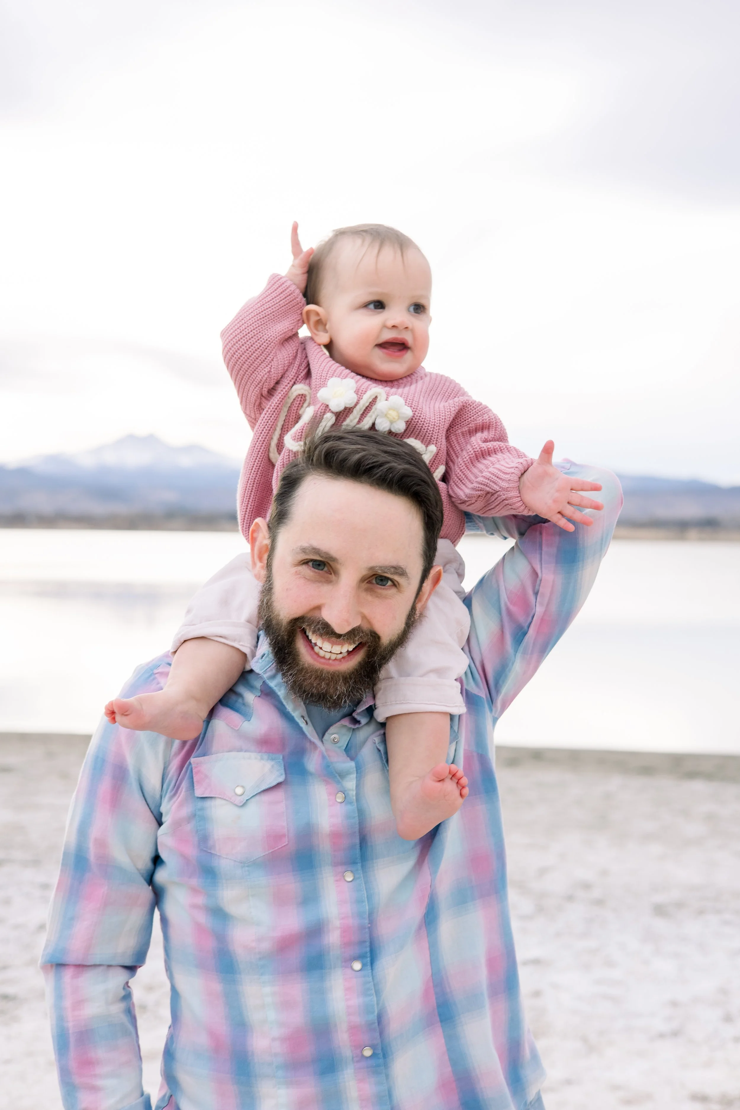 A smiling man with a beard carrying a young child on his shoulders near a lake with mountains in the background.