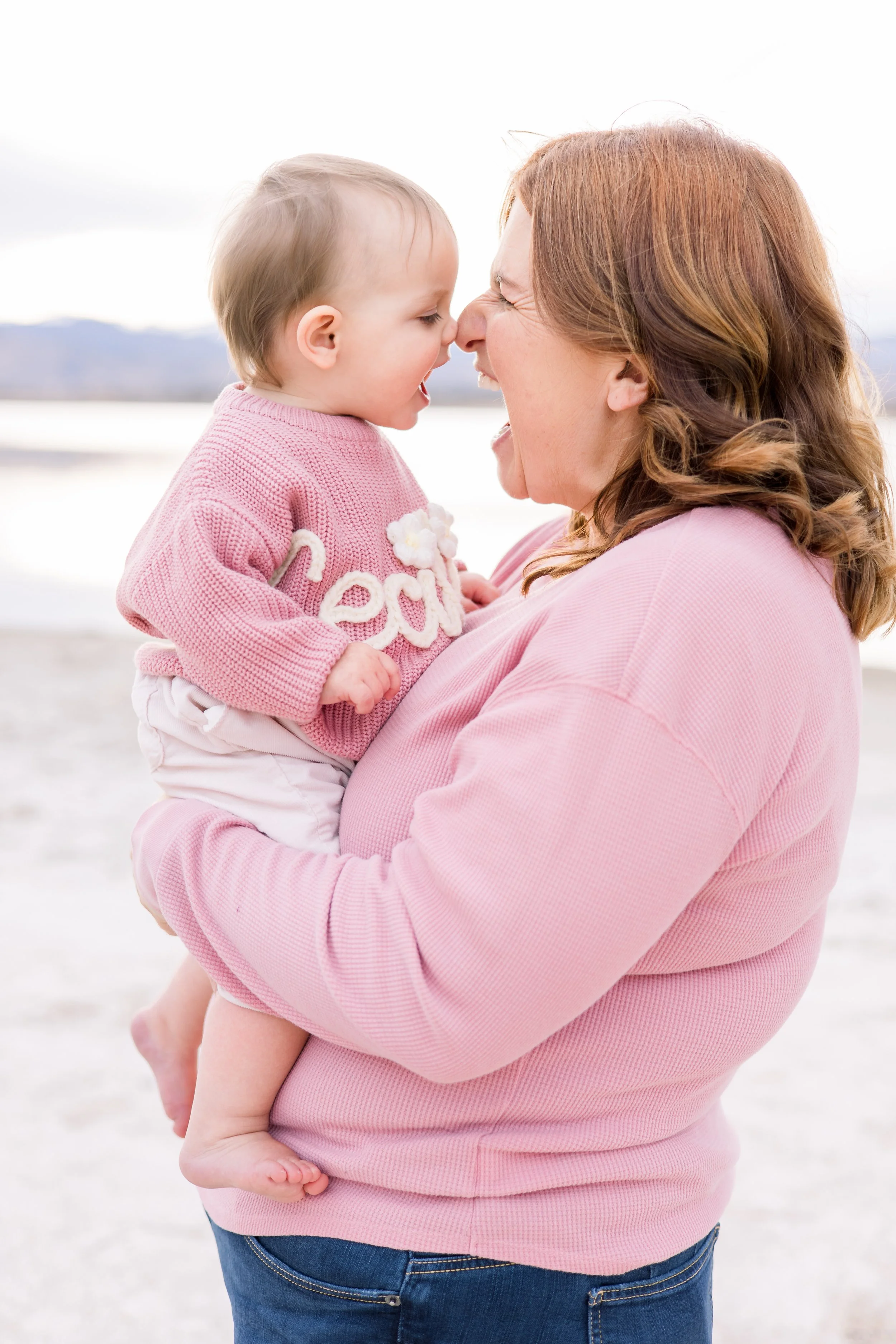 A woman and a young child are smiling and playing together outdoors near a body of water, with mountains in the background. The woman is holding the child and they are touching noses, both joyful.
