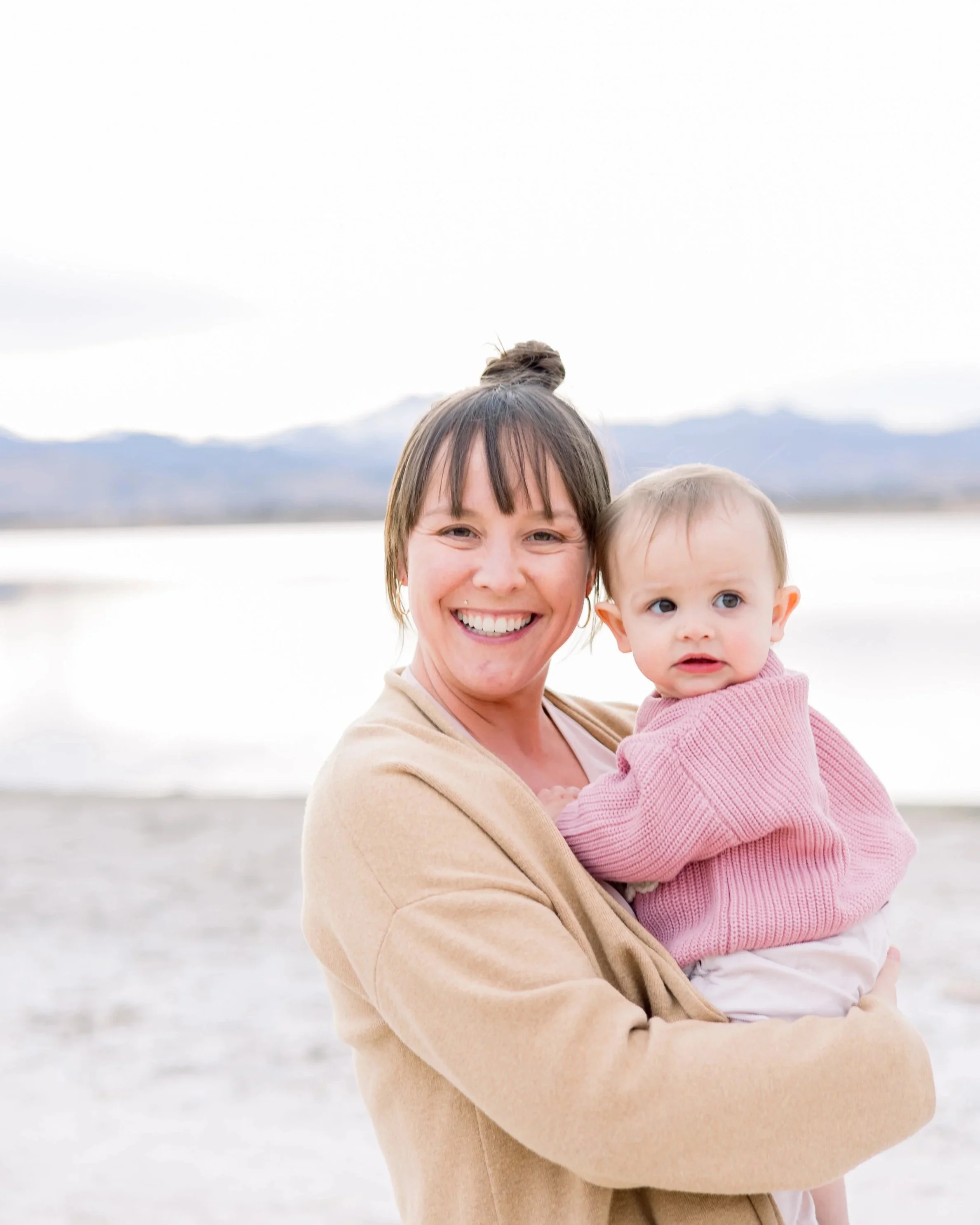 A smiling woman holding a young girl in pink sweater outdoors near a body of water, with mountains in the background.
