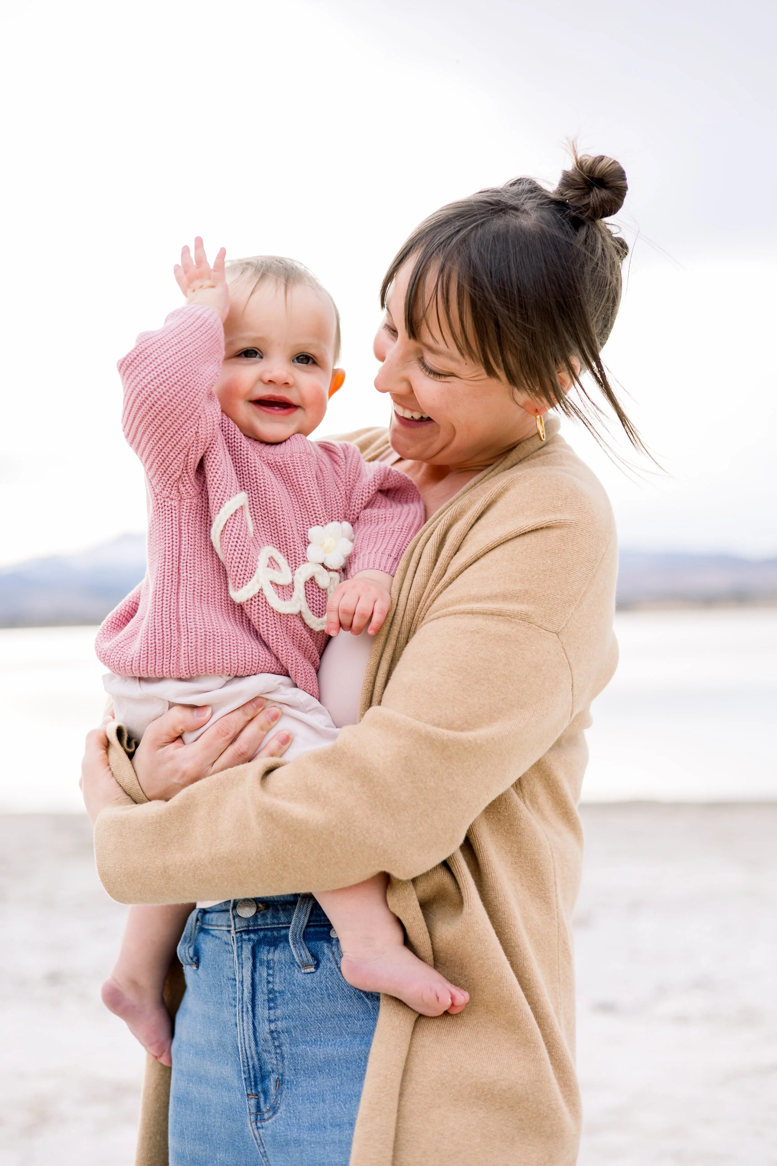 A woman holding a smiling toddler girl outdoors near a body of water.