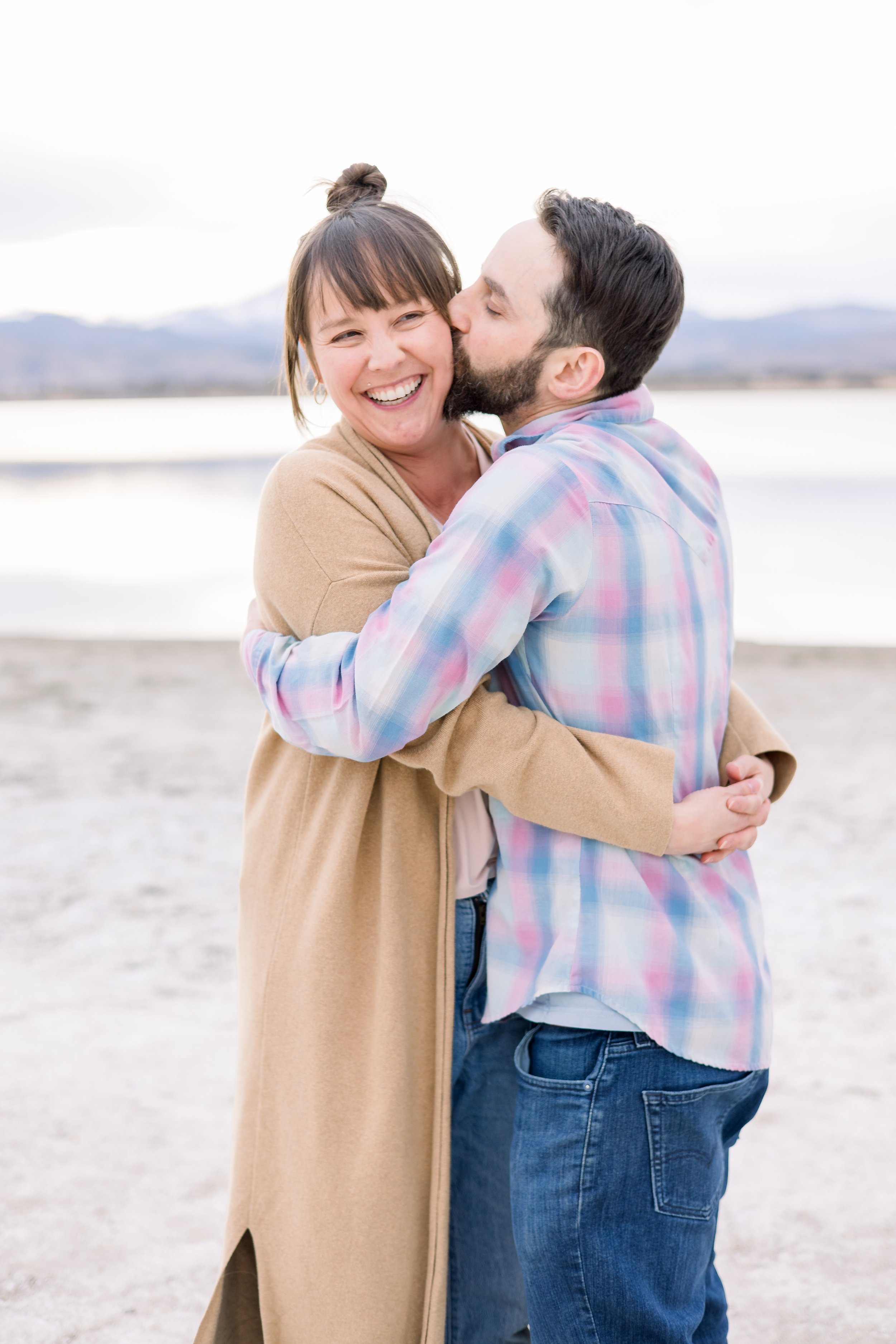 A man and woman hugging on a beach with mountains in the background. The man is kissing the woman on the cheek, and she is smiling.