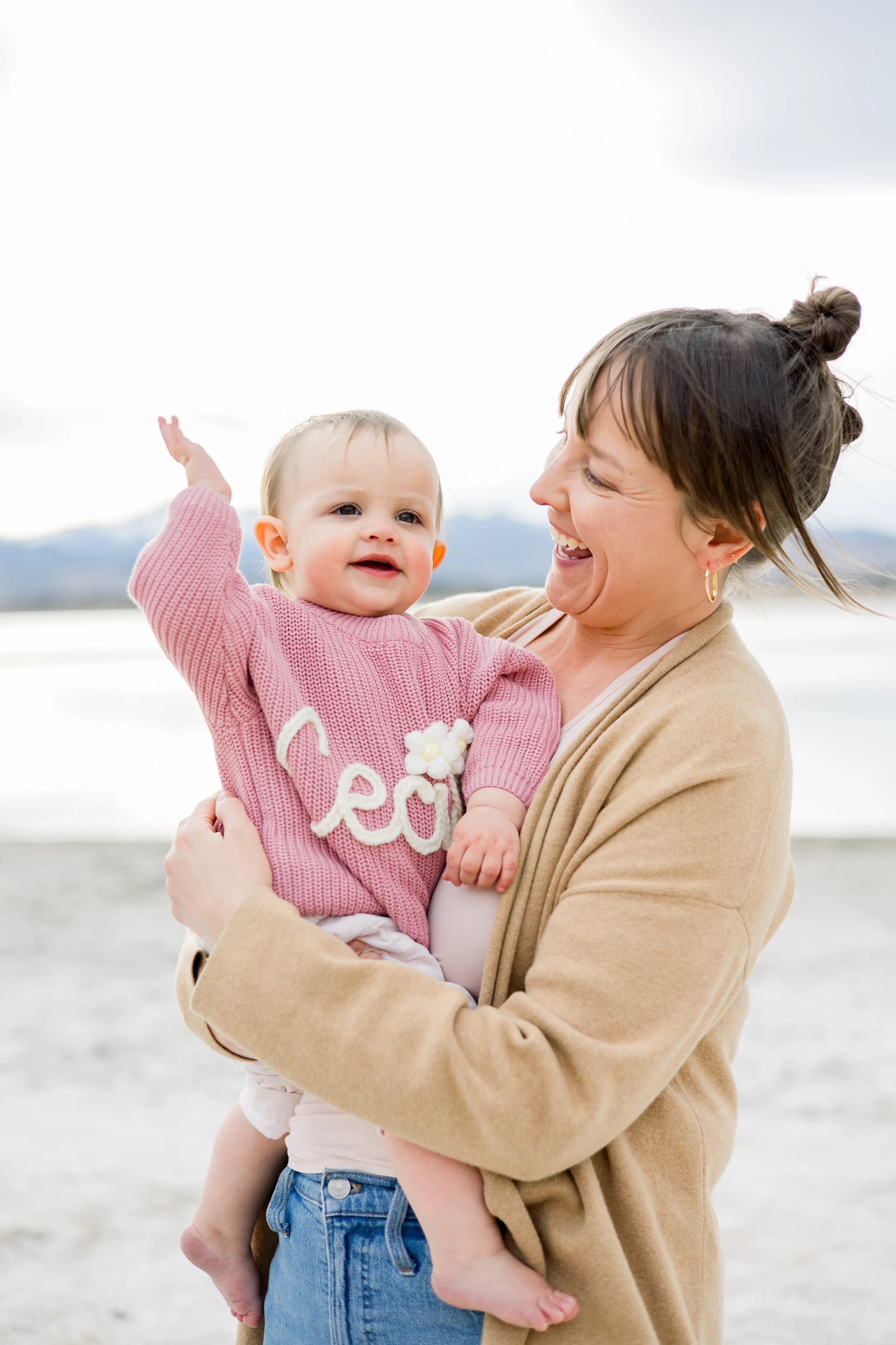 A woman holding a smiling baby girl outdoors near a body of water with mountains in the background.