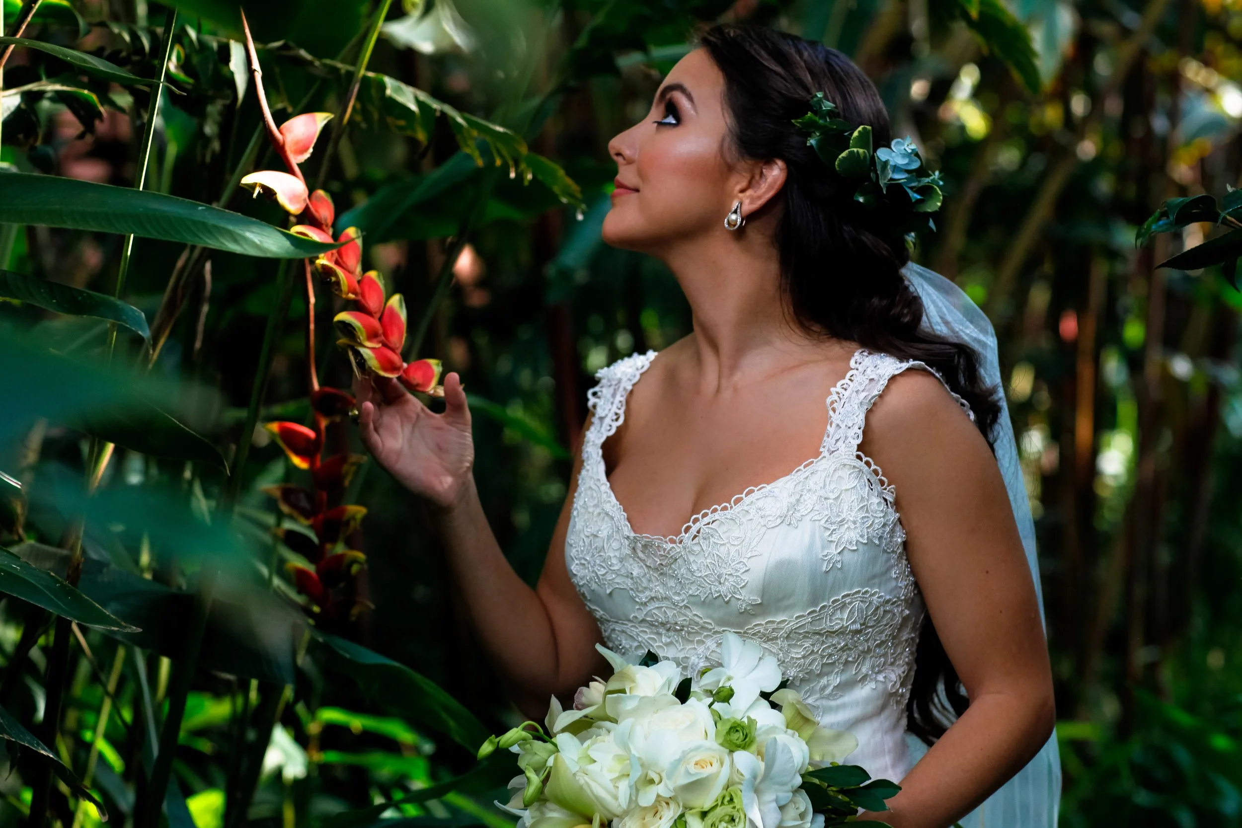 In a tropical garden, the bride gently reaches toward a vibrant bloom, captured in a quiet, intimate moment of anticipation.