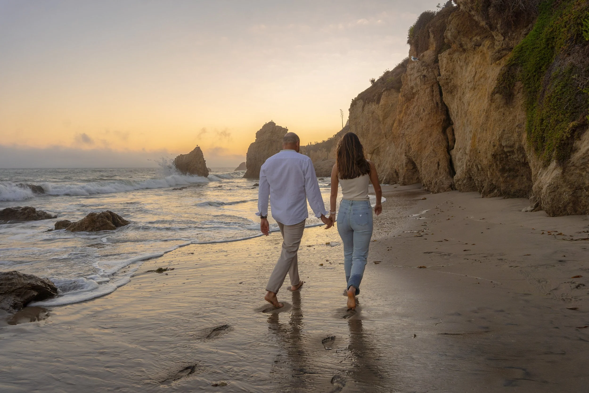 A quiet, intimate moment as the couple walks hand in hand along the shoreline at sunset, framed by golden light and rugged coastal cliffs.