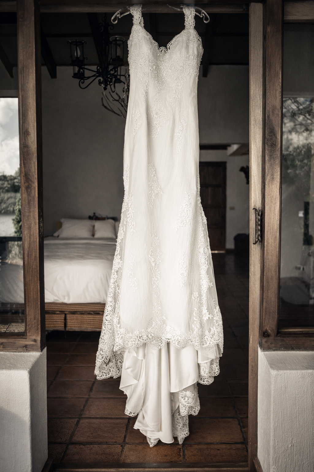 A lace wedding gown hangs in a rustic doorway, framed by warm wood tones and soft natural light, setting the tone for a timeless bridal story.