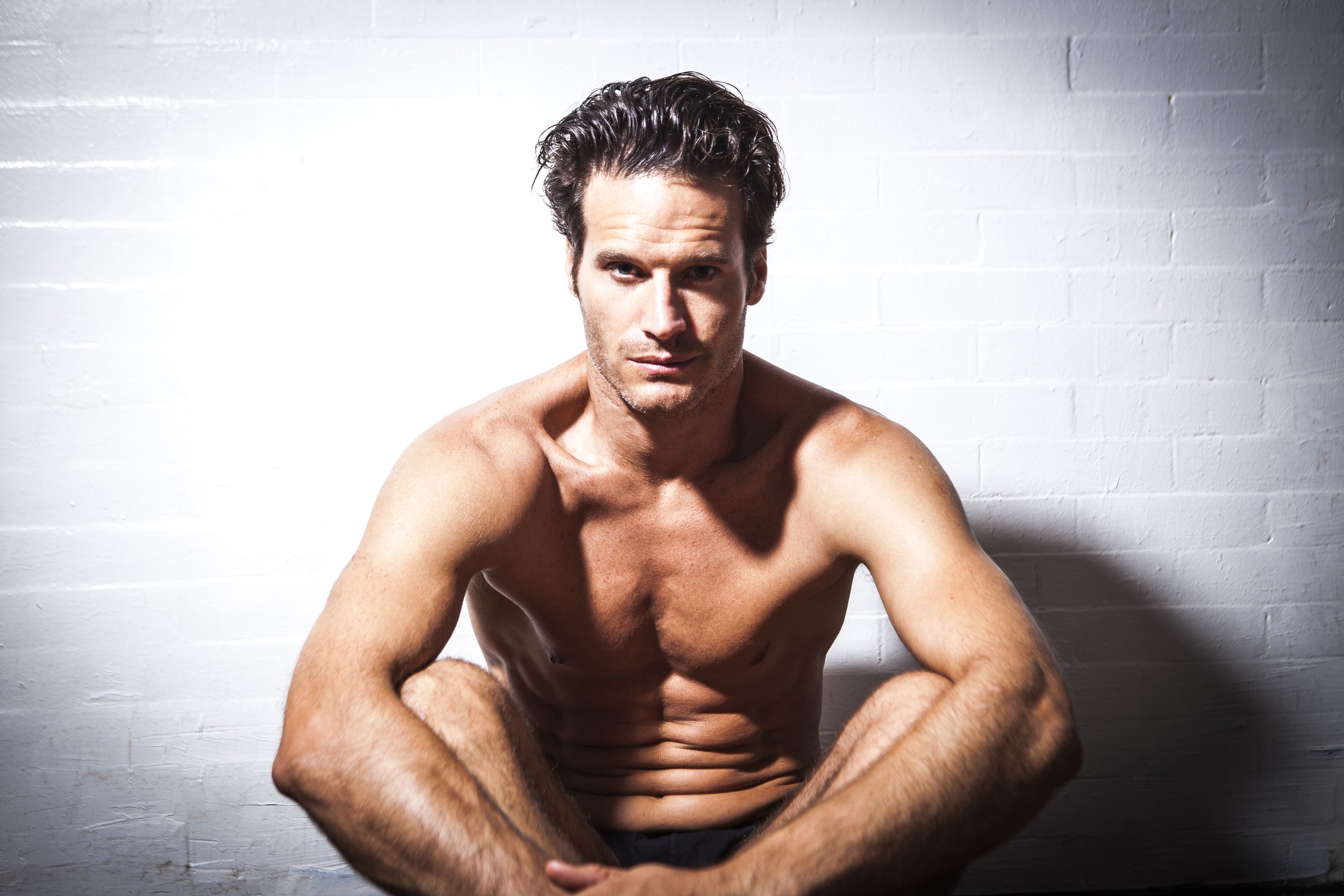 shirtless groom seated portrait against white wall.