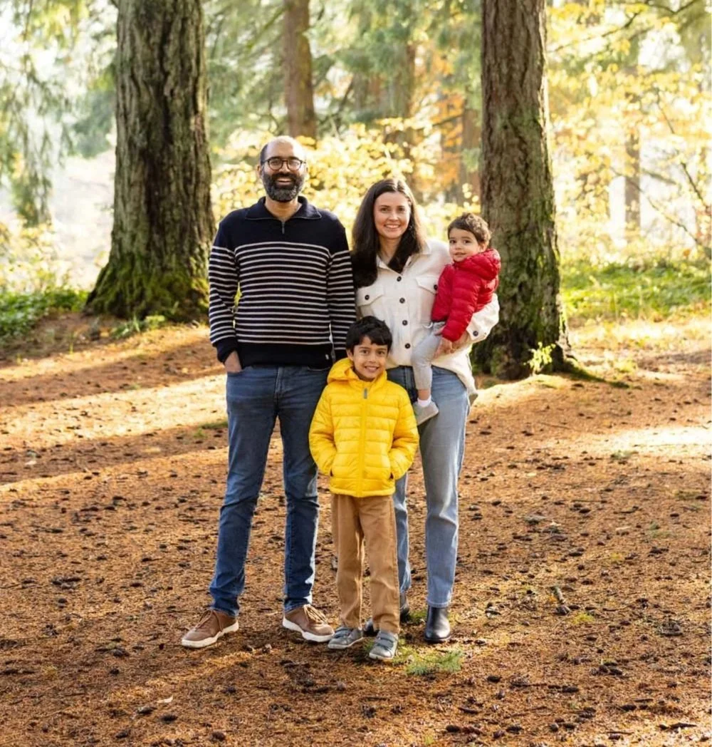 Anit and his family of four standing outdoors in a wooded area during autumn, smiling for the camera.