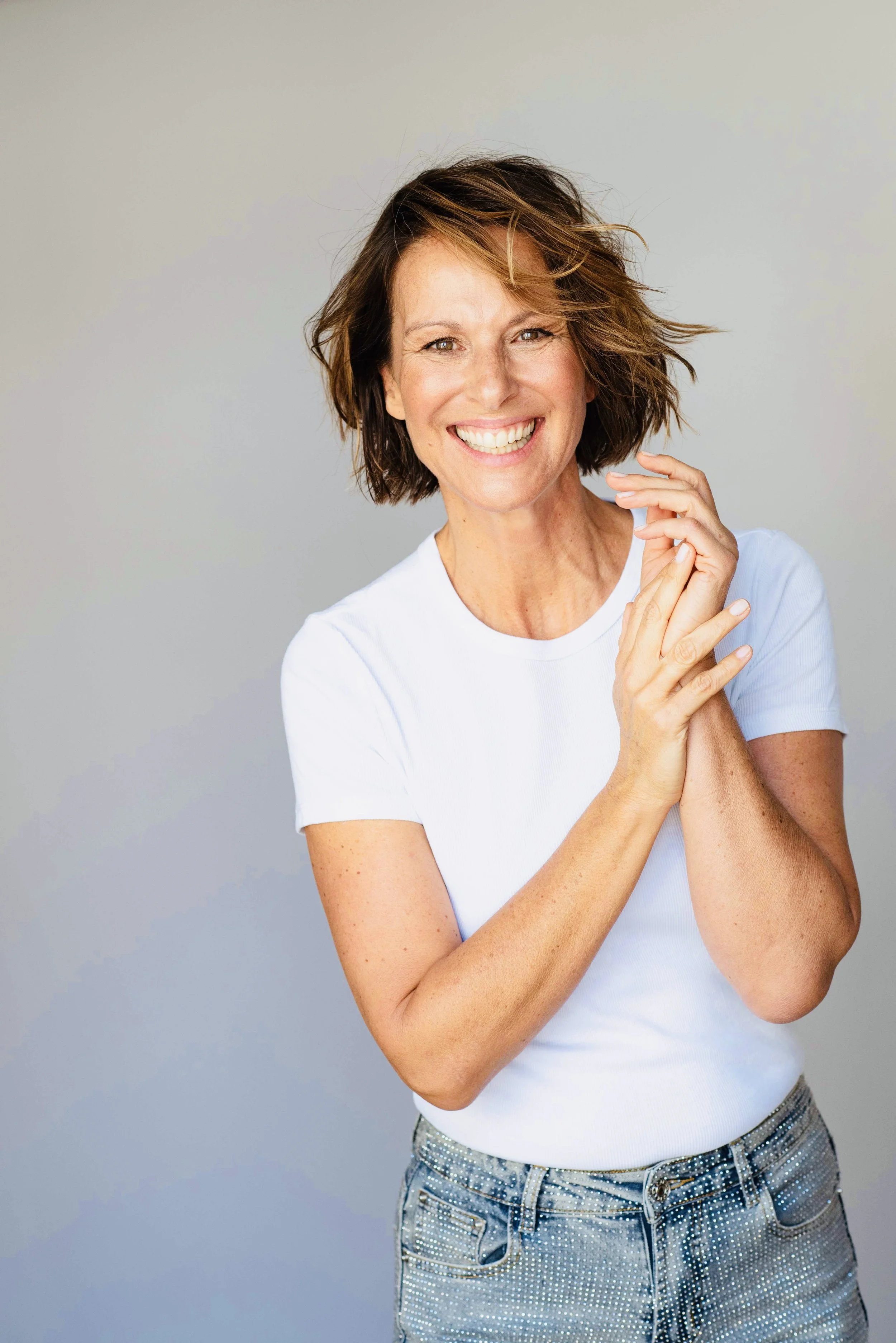 Eine lachende Frau mit braunen, gewellten Haaren trägt ein weißes T-Shirt und eine Jeanshose vor einem neutralen Hintergrund.
