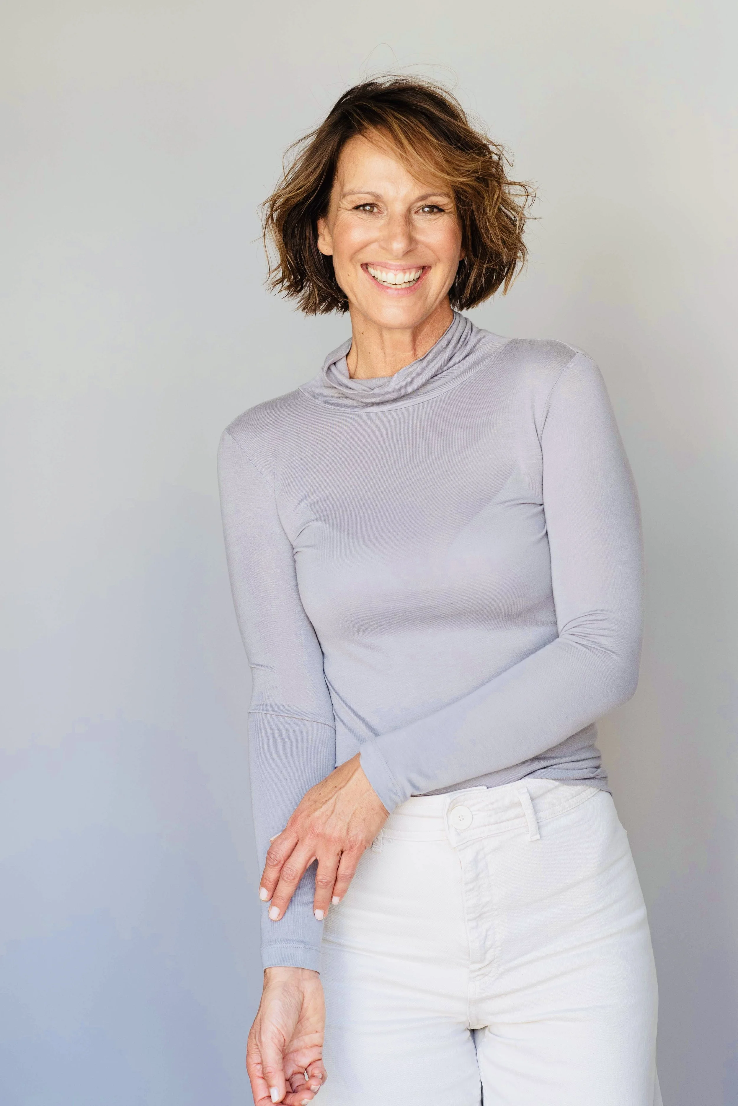 Eine lächelnde Frau mit kurzen, lockigen braunen Haaren in einem hellgrauen Pullover und weißen Hosen vor hellem Hintergrund.